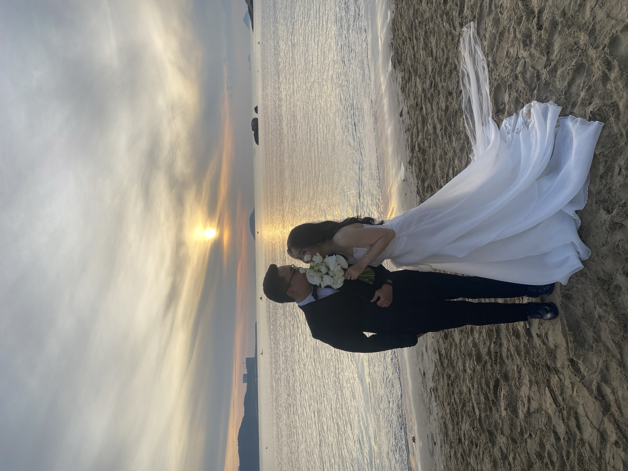 A newly married couple stands on a sandy beach in Nha Trang, Vietnam, during a sunset. The groom, wearing a black tuxedo with a bow tie, stands on the left, while the bride, in a white wedding dress with a flowing train, is on the right. The bride holds a bouquet of white flowers and leans towards the groom, who is looking at her. The sun is setting over the ocean, casting a warm glow on the water and sky. In the distance, islands and buildings are visible along the coastline. The waves gently lap at the shore, and the sand is marked with footprints.