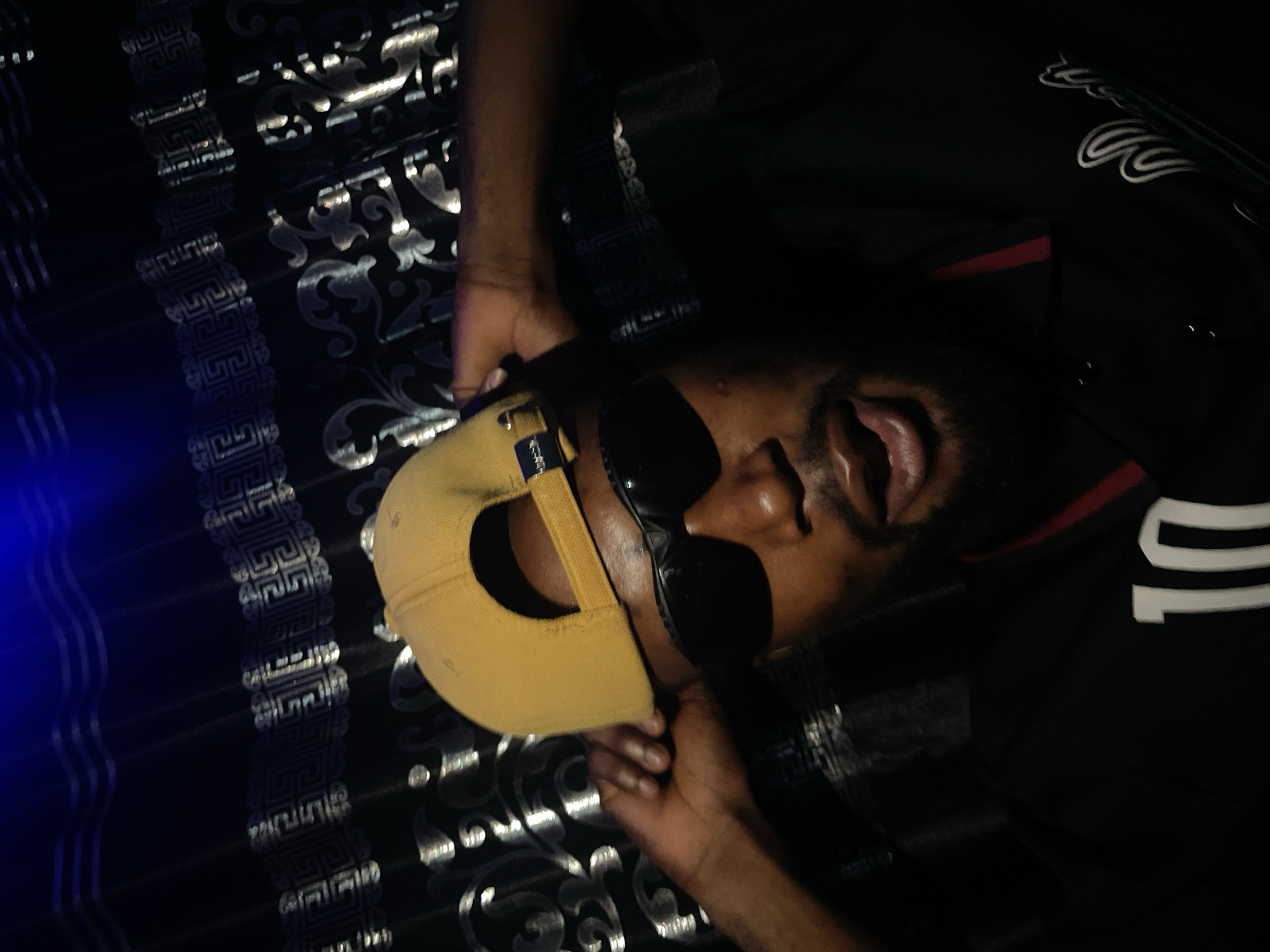 A man is lying on a dark bedspread with silver patterns. He is wearing a yellow cap, black sunglasses, and a black shirt with the number 10 in white on it. He is lying on his back with his mouth open. The location is unknown and cannot be determined from the image.