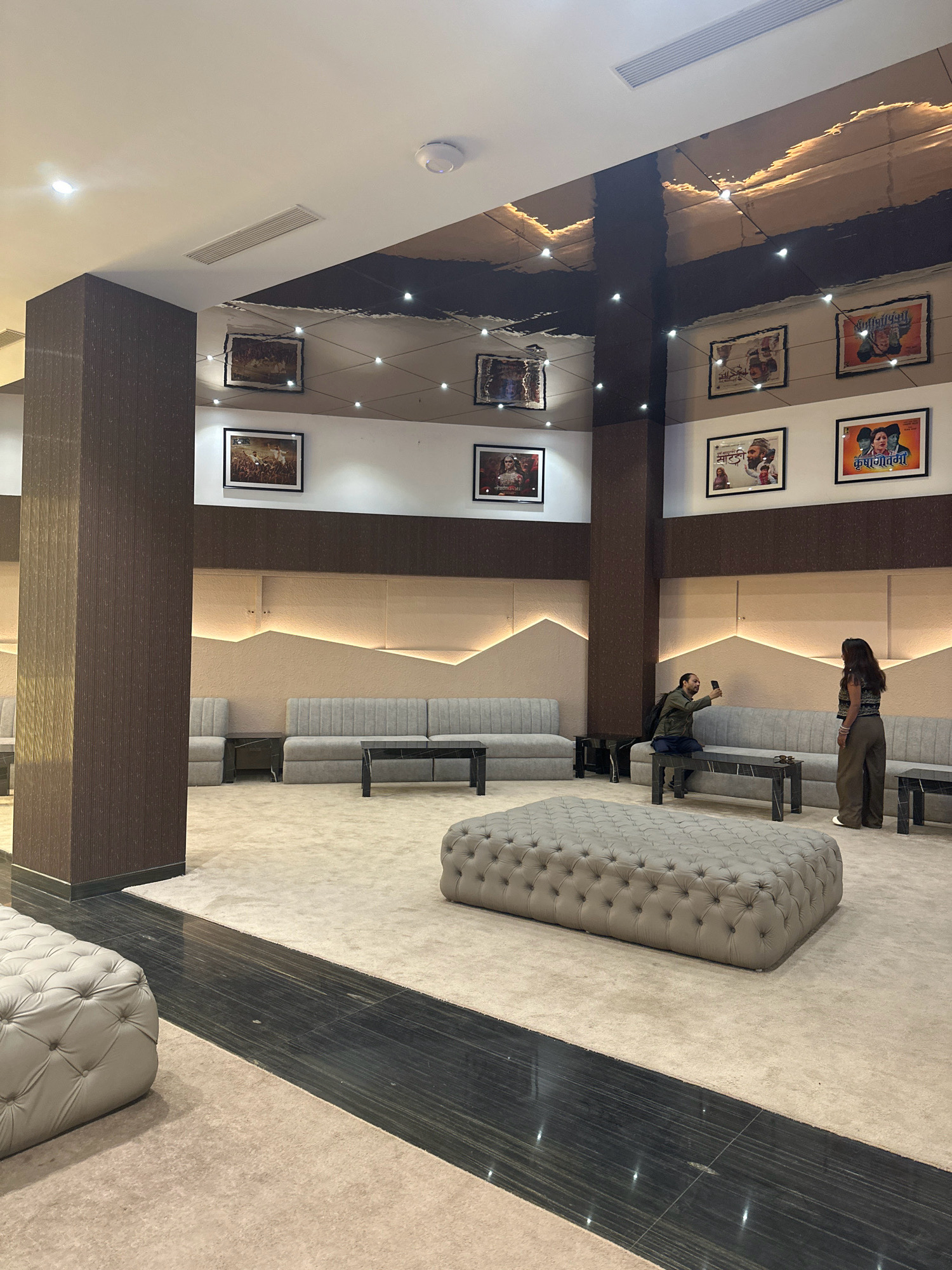 The image shows the interior of a modern waiting area or lobby, likely in Lalitpur, Nepal, possibly a cinema or entertainment venue, given the movie posters displayed on the walls. The space is furnished with sleek, upholstered seating arrangements, including long grey sofas and a large, tufted ottoman in the center of the room. The flooring is a combination of plush beige carpet and dark, reflective tile. The walls are adorned with framed movie posters, and the ceiling features a striking, reflective design with embedded lights, creating a dramatic effect.
Two individuals are present in the scene. On the left, a person is seated on a sofa, holding up a mobile phone, seemingly taking a photograph or video. On the right, another person stands near the sofa, facing the seated individual. Both are dressed casually. The overall ambiance suggests a comfortable and stylish waiting area.