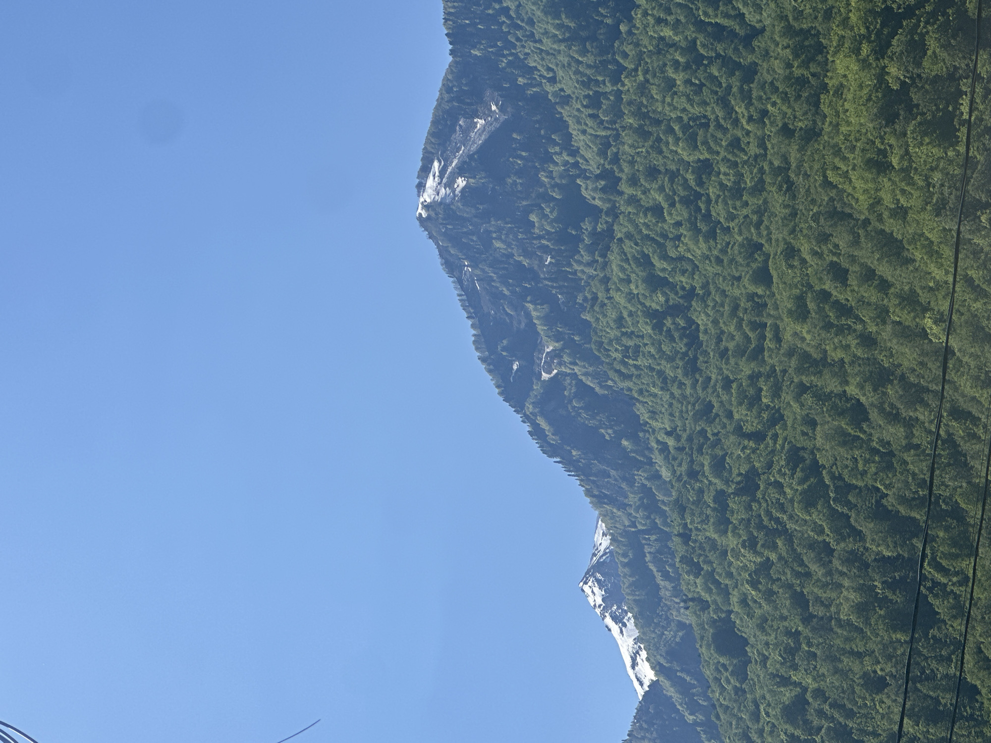 The image presents a serene mountain landscape under a bright, clear blue sky, set in Krasnaya Polyana, Russia. A large mountain range, densely covered with vibrant green trees, dominates the right side of the frame. Scattered across the rugged mountain face are several patches of white, likely snow or bare rock, contrasting with the dark green foliage. The sky, occupying the left and upper portions, is a uniform and unblemished light blue, indicating clear, sunny weather. A subtle, circular smudge, possibly a lens dust spot, is faintly visible in the upper left area of the sky. In the lower right foreground, dark, thin lines, resembling power lines or ski lift cables, are visible against the backdrop of the forested slope. The scene depicts a vast natural environment, likely captured during daytime, with no individuals or text present.