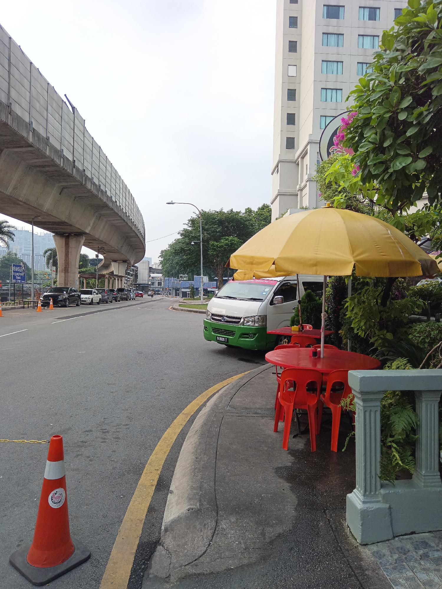 The image shows a street scene in Kuala Lumpur, Malaysia. In the foreground, a yellow umbrella shades two red tables with red chairs, set up on a sidewalk next to lush green foliage. To the left, a wet asphalt road curves away, marked by a yellow line and a prominent orange traffic cone. In the background, a multi-story building with a light-colored facade and numerous windows stands tall. An elevated concrete structure, likely part of a transportation system, arches over the road and sidewalk. Several parked cars are visible along the roadside, and a white van with green accents is positioned near the tables and chairs. The sky is overcast.