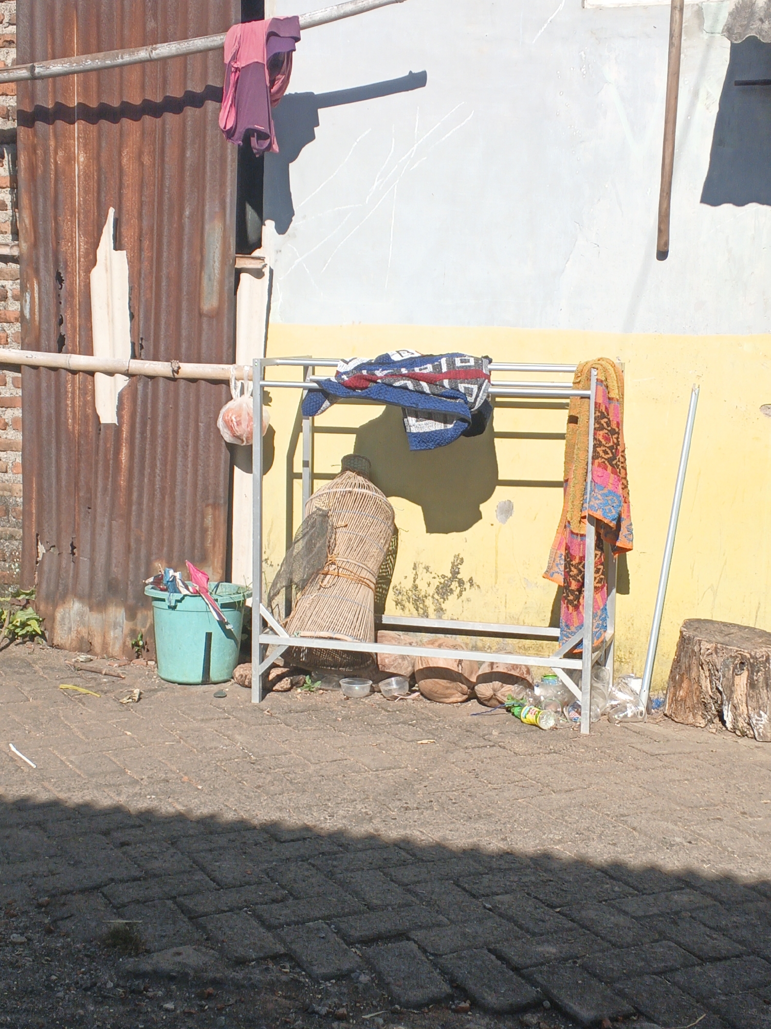 In Singolatren, Indonesia, an outdoor scene unfolds with a focus on laundry and discarded items. A corrugated metal wall, weathered and rusty, forms a backdrop. A metal drying rack stands centrally, holding a blue and red patterned blanket and an orange and pink patterned towel. To the left, a green plastic bucket contains pink and red clothing. Behind the bucket, a woven birdcage rests on the ground. Several plastic bottles and other debris are scattered near the base of the drying rack, alongside a rough-hewn wooden log on the right side. The ground is paved with interlocking stones. The scene is illuminated by strong sunlight, casting distinct shadows.