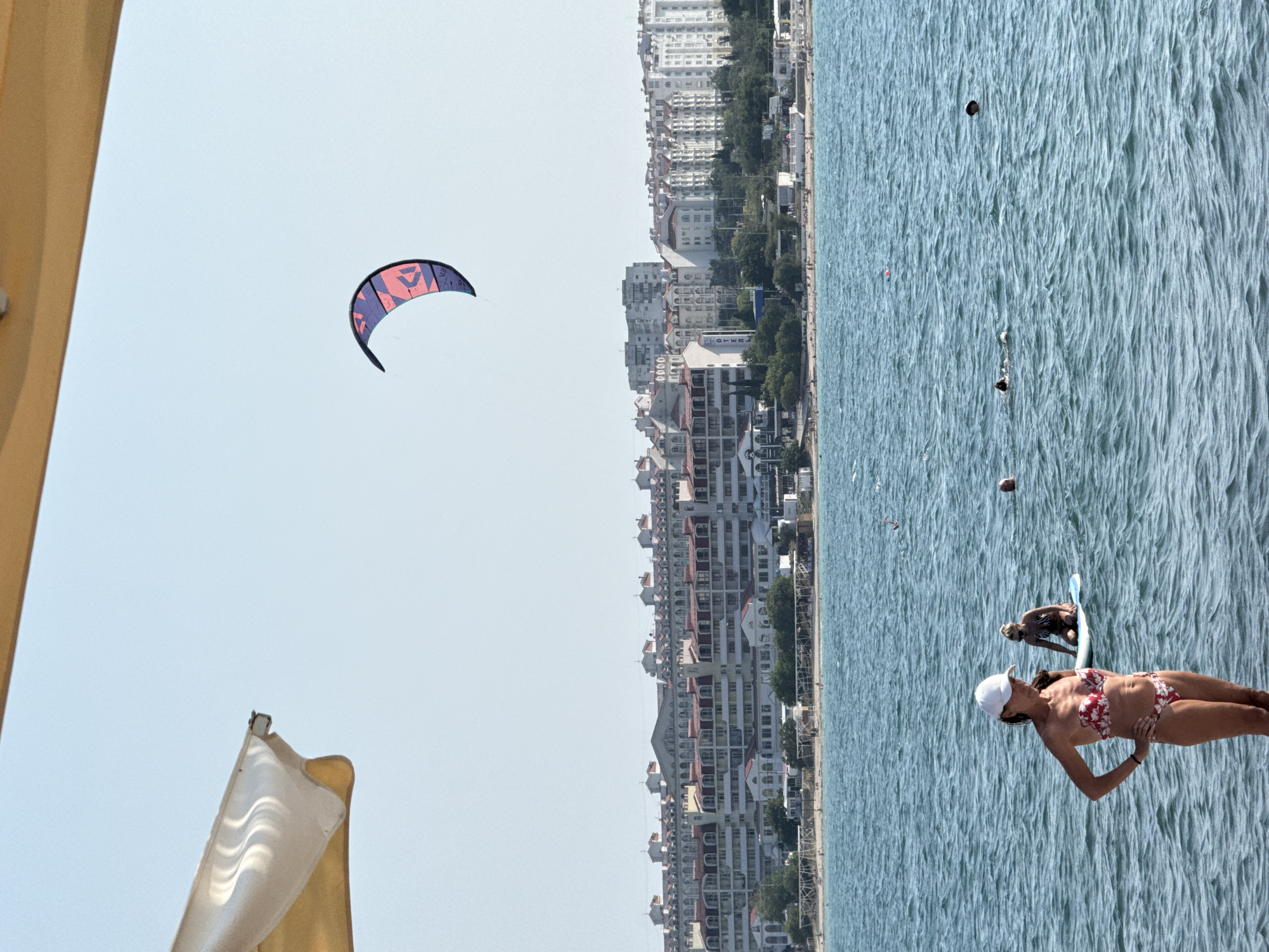 An outdoor, eye-level, medium shot shows a beach scene under a clear sky with a kiteboarder in the distance. The foreground is dominated by the turquoise blue, choppy water, with a woman in a red and white floral bikini and white baseball cap standing with her hands on her hips. Behind her, to the right, another woman in a striped bikini kneels on a blue paddleboard. In the background, a long stretch of white and red-roofed buildings lines the coast, with trees and beach umbrellas visible on the shore. A large, purple and pink kite is flying in the sky above the water. The edge of a yellow umbrella and a white awning are visible in the upper left of the frame.