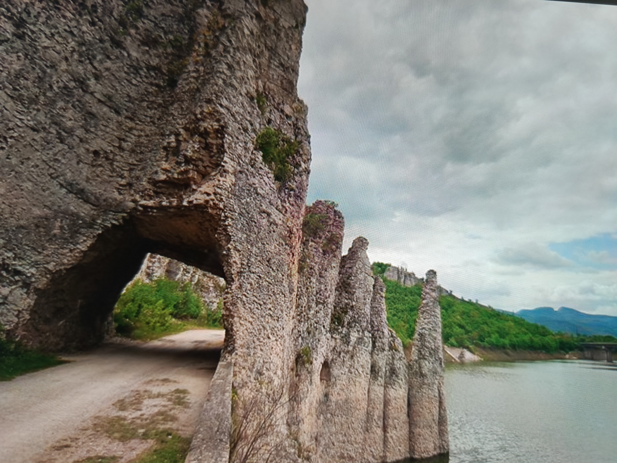 This media file captures a natural landscape with a rock formation that has been sculpted by erosion. The foreground features a rough, textured rock face, with a large archway carved into it, revealing a dirt path leading through. The path is lined with greenery on either side. To the right of the rock formation, several tall, slender rock pillars rise from the ground, also showing signs of erosion and weathering.
Beyond the rock formations, a body of water stretches towards the horizon, with a lush green hillside rising from its edge. The hillside is covered in trees and vegetation, with more distant mountains visible under a cloudy sky. A bridge or structure can be seen in the distance, crossing the water.
The scene appears to be set during the daytime, with overcast weather indicated by the gray, diffused light and cloudy sky. The location is Varna, Bulgaria. There are no people or text visible in the image. The overall mood of the image is serene and natural, highlighting the power of geological processes and the beauty of the natural world.