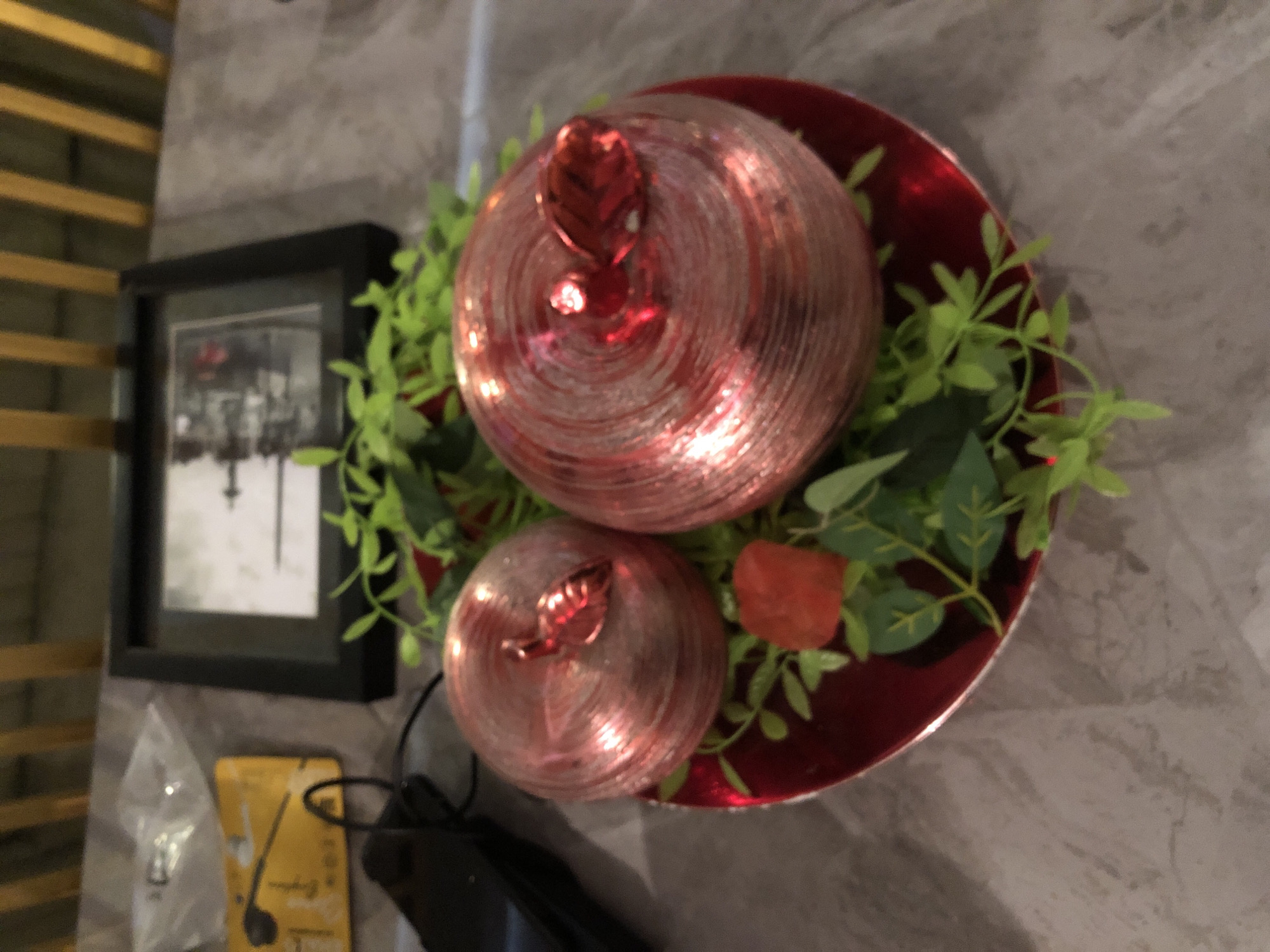 The image captures an indoor still life arrangement, primarily featuring a decorative centerpiece. At the heart of the composition are two spherical objects, resembling apples or pomegranates, showcasing a shimmering, textured finish in shades of pink and red. Each sphere is topped with a small red, leaf-like stem. The larger sphere is positioned behind the smaller one. These decorative elements rest on a glossy, dark red round plate or shallow bowl, which serves as their base. Artificial green foliage, appearing as small sprigs with light green leaves, is arranged around the spheres on the red plate. A small, solid red decorative item, possibly an artificial berry or fruit, is also visible among the green leaves.
To the left of this central display, a black-framed picture or artwork is placed vertically. The image within the frame is black and white, possibly depicting a city scene or architectural details, though specific elements are indistinct. Below the framed artwork, on the grey surface, a clear plastic bag with faint text, potentially "LD," is visible. Next to it is a small, rectangular yellow box, which appears to be packaging for "Earphones," as suggested by a visible graphic of an earbud and partial text. Black cables or wires are also present on the surface.
All objects are arranged on a flat surface, likely a table or shelf, characterized by a grey, marble-like or textured finish. In the upper left background, a structure with horizontal wooden slats, possibly blinds or wall paneling, is partially visible. The scene is static, depicting a curated decorative display in an indoor setting.