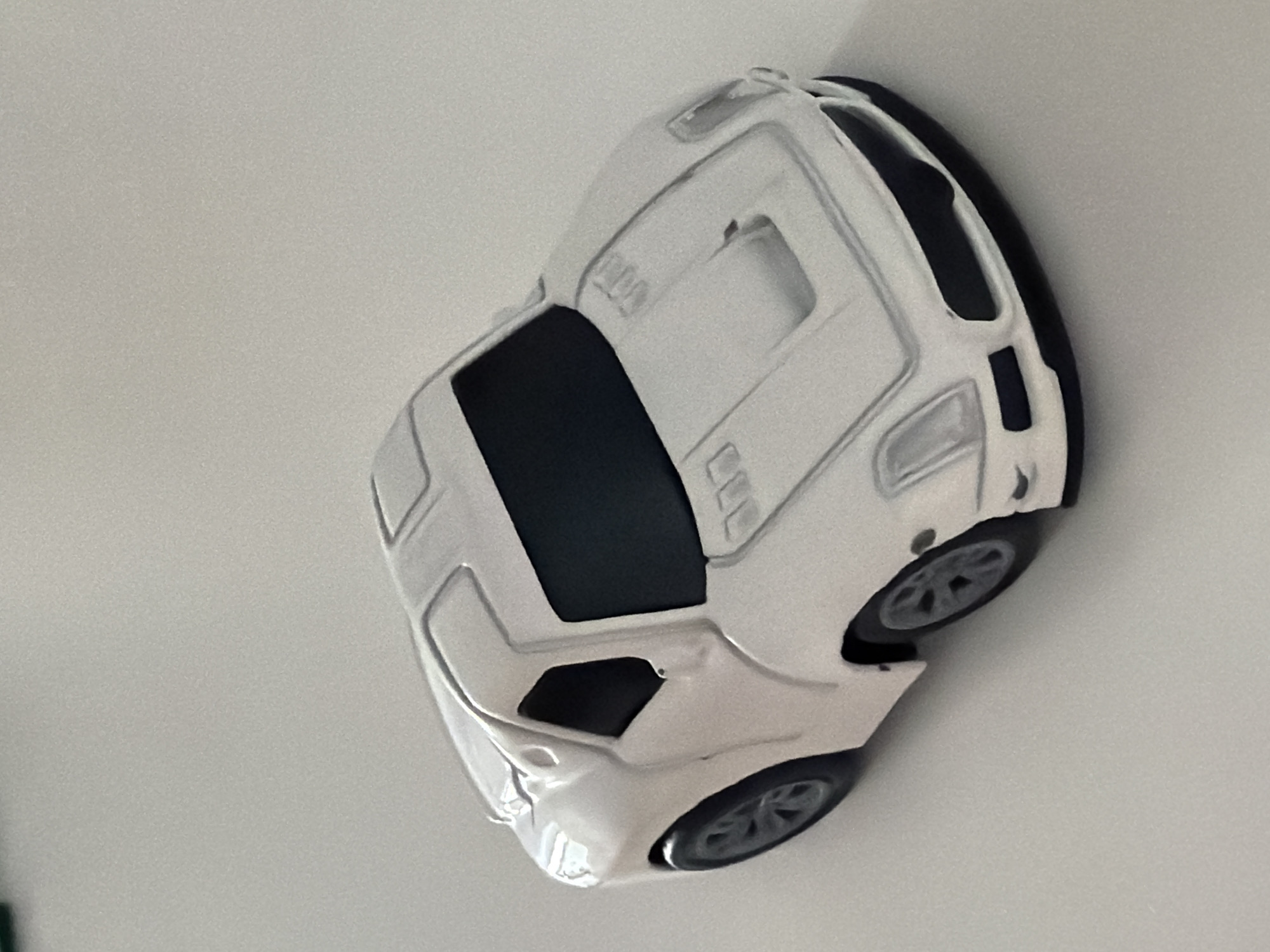 The image shows a close-up, top-down view of a white toy car with black accents, possibly a miniature sports car or a futuristic vehicle. The car is positioned diagonally against a plain, light-colored background, which appears to be a wall or a tabletop. The lighting suggests an indoor environment, likely with diffused light, as there are no harsh shadows. The car has detailed features, including visible wheels with intricate rims, a blacked-out window area, and sculpted body lines that suggest aerodynamic design. The overall impression is of a static object, likely a collectible or a child's toy, presented in a clean and minimalist setting. There is no visible text in the image. The context of Łódź, Poland, does not provide specific visual cues within the image itself but situates the object within a geographical location.