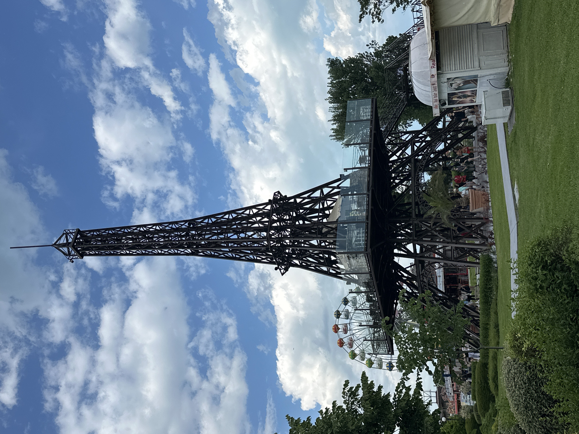 The image shows a replica of the Eiffel Tower in Varna, Bulgaria, set against a partly cloudy sky. The tower is made of dark metal lattice and features a glass-enclosed section. A Ferris wheel is visible near the base of the tower. Green trees and bushes are present in the foreground. There is also a white concession stand with a sign that says “Real Italian Gelato” and people standing nearby. A paved path is adjacent to the stand.