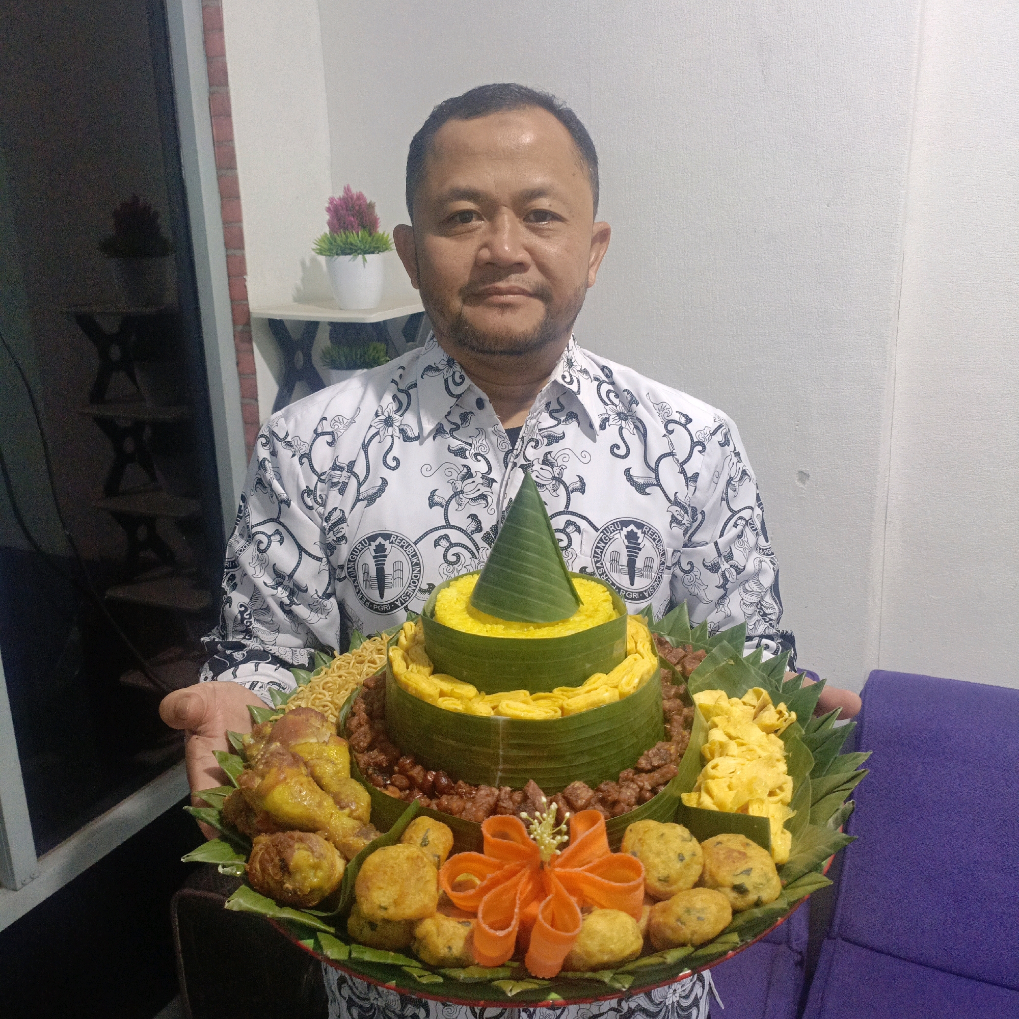 A man with short dark hair and a goatee, wearing a white patterned shirt with a dark emblem on the left chest, holds a tiered food offering in front of him. The offering, presented on a circular platter lined with banana leaves, is topped with a cone made of banana leaves, filled with yellow rice. The tiers of the offering are decorated with more banana leaves, and include noodles, fried chicken pieces, fried tempeh, and small fried balls. An orange carrot decoration resembling a flower sits in the center of the bottom tier. The man is standing indoors, with a wall behind him and a dark, partially visible object to his left. The lighting is even, and the focus is on the man and the food. This scene is likely from Salatiga, Indonesia, given the man's attire and the traditional Indonesian food presentation.