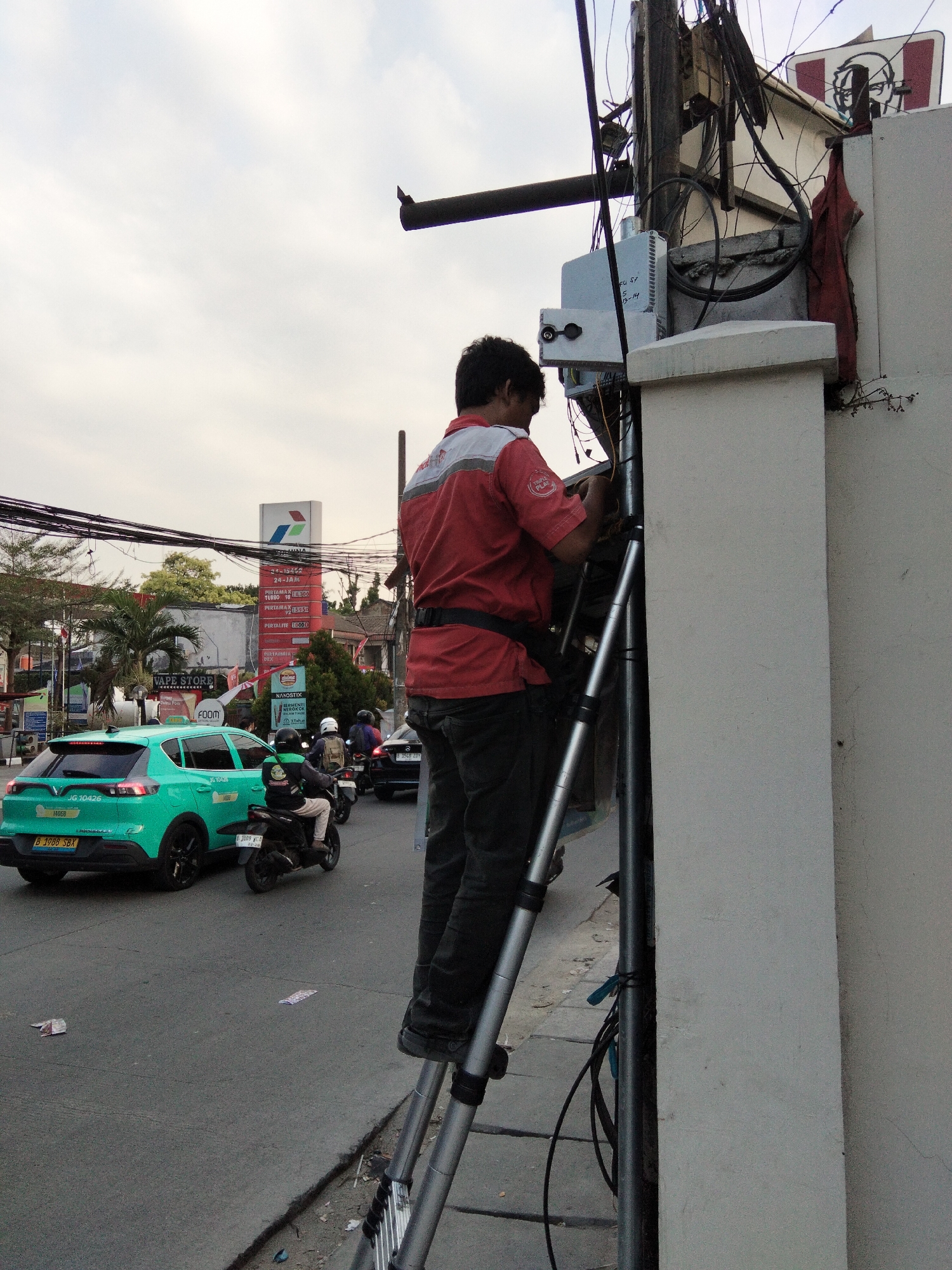 The image depicts a man in a red shirt and dark pants standing on a ladder, working on electrical equipment mounted on a pole near a white wall. The man is facing away from the camera, with his hands engaged with the equipment. In the background, there is a street with traffic, including a green car and several motorcycles. A Pertamina gas station sign is visible, indicating the likely location as a commercial area in South Tangerang, Indonesia. The sky is overcast.