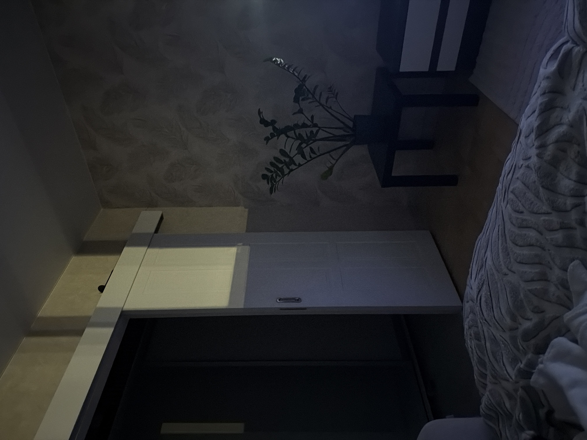 The image depicts a dimly lit bedroom. In the foreground, a textured duvet covers a bed. To the left, a sliding door to a closet is partially open, revealing a dark interior. The wall to the right features a textured wallpaper with a pattern of swirling strokes in muted tones. A small, dark square table stands against this wall, topped with a potted plant. To the right of the table, a white nightstand with two drawers is visible, partially obscured by shadow. The overall lighting suggests nighttime or low artificial light. The scene is consistent with an interior residential space in Izhevsk, Russia.