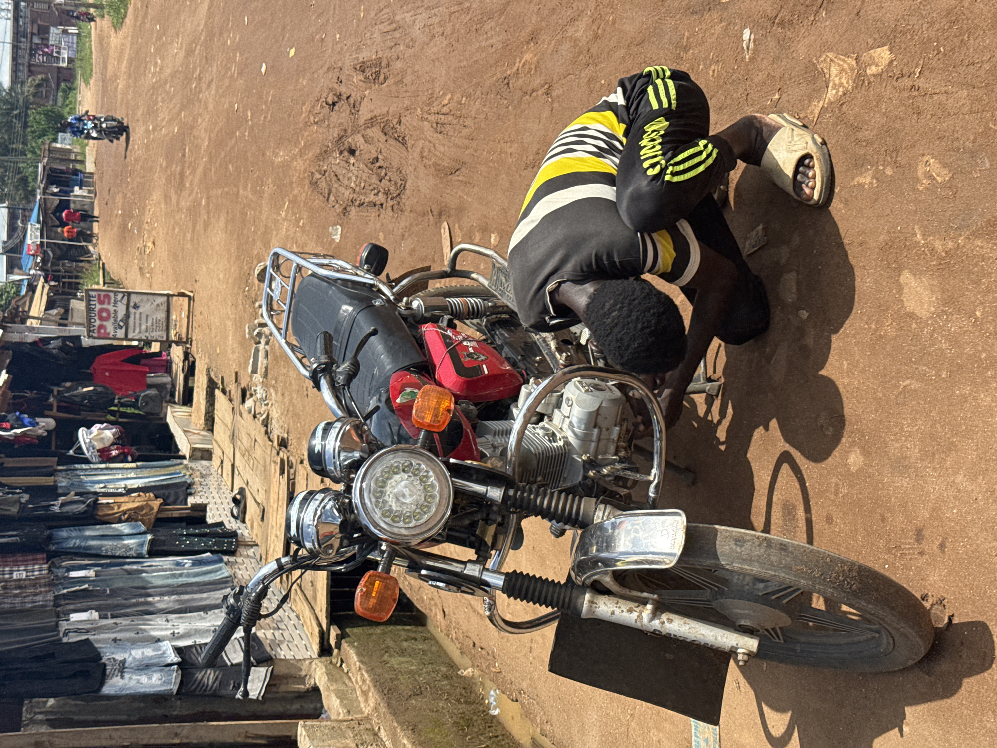 The image captures a vibrant outdoor scene on a sunny day in Ihiagwa, Nigeria, depicting a common roadside activity. The main subject is a man, dressed in a distinctive black, yellow, and white striped shirt and black pants with yellow stripes that bear text resembling "SPORTS." He is focused on the front section of a parked motorcycle, bent over as if inspecting or repairing it. He wears light-colored sandals on his feet.
The motorcycle is a prominent feature, primarily black and red, with numerous chrome details including the handlebars, engine components, and the casing around its round LED-style headlight and orange turn signals. The bike's front wheel rests directly on the unpaved, reddish-brown dirt ground.
In the background, the scene extends along the dirt road, revealing more of the local environment. To the left, a series of makeshift market stalls or shops display various goods, predominantly stacks of folded clothing like jeans and shirts. Further down the road, other individuals and a motorcycle can be seen, indicating a degree of public activity. A visible white sign with a red top prominently advertises "FAVOURITE POS" and "Available Here," with smaller print detailing services such as "Cash Deposit," "Cash Withdrawals," and "Bills Payment." The strong, clear shadows cast by the man and the motorcycle indicate bright daylight, likely mid-day, under clear skies. The overall impression is one of everyday life and commerce in a bustling, unpaved street setting.