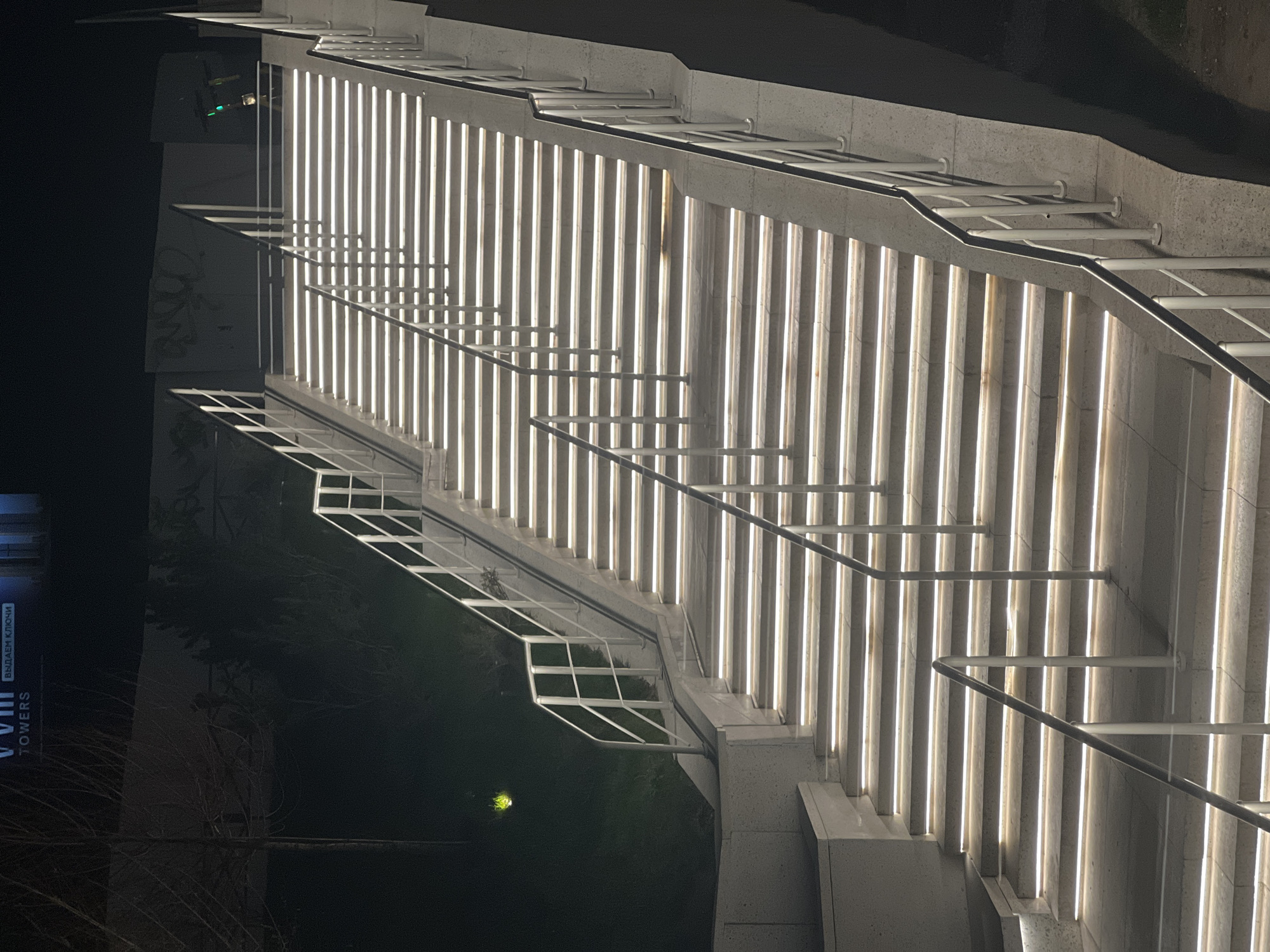 The image displays a modern, brightly illuminated outdoor staircase or ramp structure at night. The photograph is rotated 90 degrees clockwise, presenting the steps horizontally across the frame, viewed from a low angle looking upwards.
The primary subjects are the wide concrete stairs, each riser of which features a prominent, cool white vertical strip of light, creating a strong, repeating linear pattern. These integrated lights are the main source of illumination for the scene. Accompanying the stairs are sleek, minimalist metal railings, composed of vertical pickets and horizontal handrails, running along the ascent. The stairs and supporting walls appear to be constructed from light-colored concrete or stone.
No people are visible, indicating no immediate activity or event taking place. The scene is static and quiet.
Notable details include the clear nighttime setting, evident from the dark background and the reliance on artificial lighting. In the dark background to the left (top-left in the rotated image), a large sign is partially visible. It clearly displays the English text "VIII TOWERS" and, above it, the Cyrillic text "ВЫДАЕМ КЛЮЧИ," which translates to "WE ISSUE KEYS." This suggests the structure is part of a building complex. A faint green light source can be seen in the distant background to the right (bottom-right). The architectural style is contemporary, highlighting functionality and illumination. The location context is Khimki, Russia.