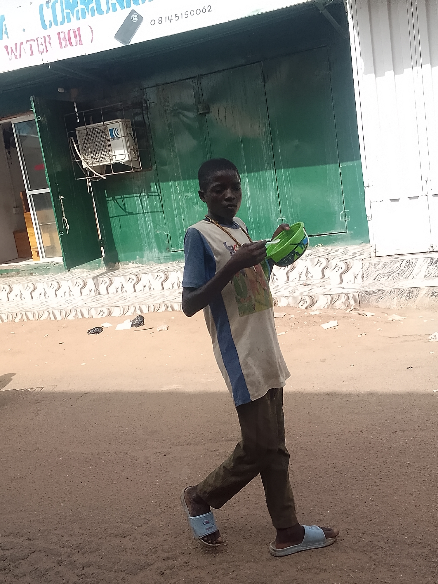 A young boy walks on a street. He wears a tan shirt with blue trim, brown pants, and blue sandals. He is holding a green bowl and a spoon. The background consists of buildings, one with a sign that reads "COMMUNI" and a phone number: 08145150062. The location is in Gombe, Nigeria.