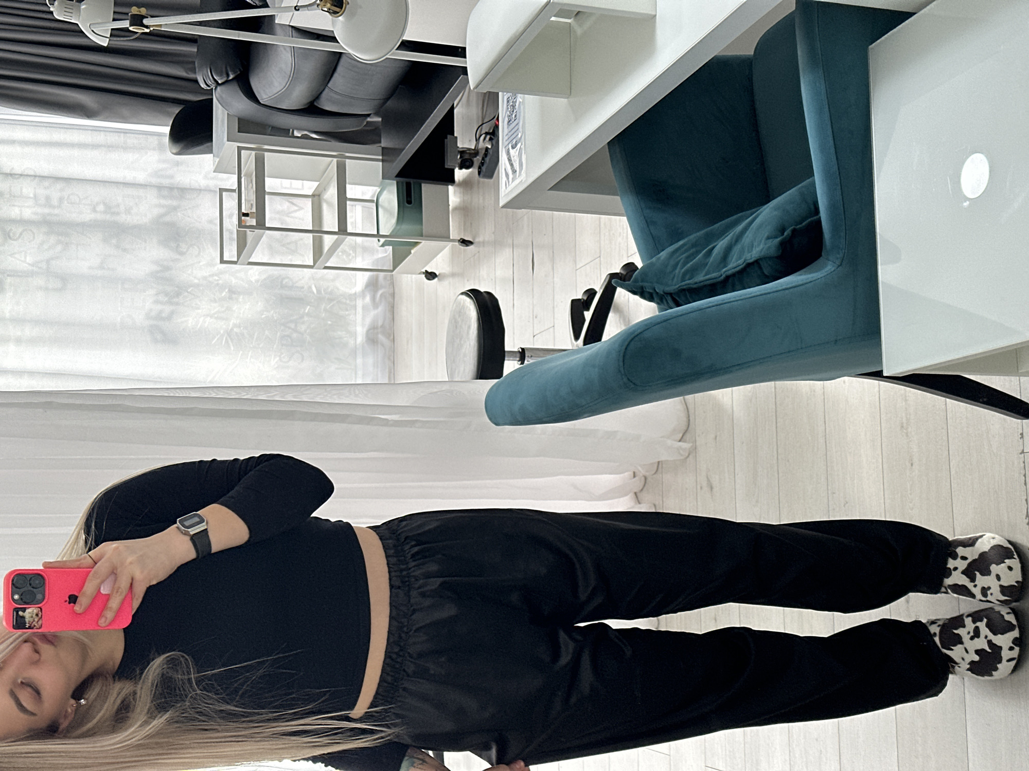 A person is standing in a room. The person is wearing a black long-sleeved shirt, black pants, and cow-print slippers. The person is holding a pink iPhone in their left hand, and is wearing a watch on their left wrist. The person has long, blonde hair. The background of the image includes a white desk, a blue velvet chair, a white bookshelf, white curtains, and a partially visible black office chair. The floor is light-colored wood. The location is likely indoors in a room. The city of Budva, Montenegro, cannot be confirmed based on the details in the image.