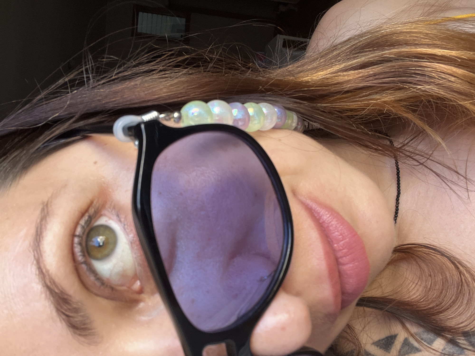 A close-up, rotated portrait shot captures a person's upper face, hair, and shoulder, likely taken in Barcelona, Spain. The subject is a person, likely female, with long, auburn-brown hair. Their face is partially obscured by a pair of black-framed sunglasses with purple-tinted lenses, which are held up, covering part of their nose and mouth. One eye, a light green or hazel color with a visible pupil and a reflection of a light source, is clearly visible and looking directly into the camera. The eyebrow above is dark and well-defined.
The person's lips are full and a rosy pink color. Around their wrist, a bracelet made of iridescent, pastel-colored beads (white, light green, purple) is visible. A thin, black beaded necklace rests on their skin near the shoulder/neck area. Further down on the shoulder/chest, a tattoo with geometric patterns is faintly visible. The lighting is soft and ambient, suggesting daytime indoors or in a shaded area, without harsh shadows. The image's rotation creates an unusual and intimate perspective, focusing on the details of the face and accessories. No visible text is present in the image.