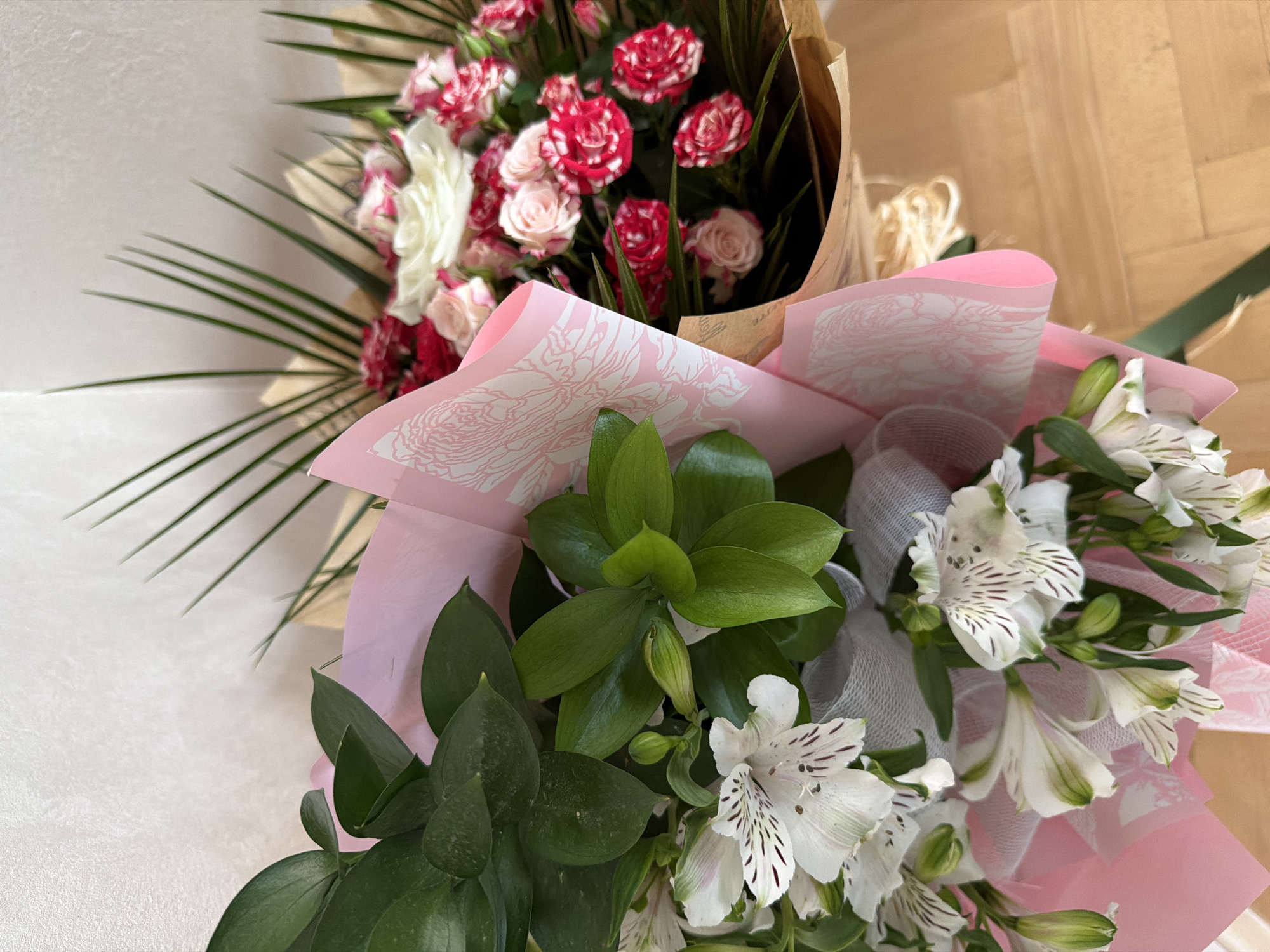 The image captures a close-up view of two distinct bouquets of fresh flowers, arranged in an indoor setting.
In the background and positioned to the left, one bouquet features a dense cluster of small blooms, predominantly roses or carnations, in shades of vibrant red, soft pink, and white, some exhibiting a speckled or marbled petal pattern. This bouquet is wrapped in plain, natural-toned brown paper and is accented with several long, slender, dark green palm-like leaves that extend outwards.
The foreground and right side of the image showcase a second bouquet, which primarily consists of white Alstroemeria (Peruvian lilies). These flowers have delicate white petals, many of which are adorned with distinctive dark streaks or spots. This bouquet is enriched with robust, broad green leaves and is elegantly wrapped in light pink paper, which is patterned with an intricate white floral design. A white, semi-transparent mesh ribbon is also visible, contributing to the arrangement.
The overall lighting is soft and even, suggesting a well-lit indoor environment. The background shows a combination of a light-colored, textured surface on the left and a warmer, possibly wooden, surface on the right. There is no specific activity taking place, and no discernible human subjects. While the location context is provided as Plovdiv, Bulgaria, there are no explicit visual cues within the image itself to confirm this. No legible text is visible on any part of the bouquets or their wrapping.