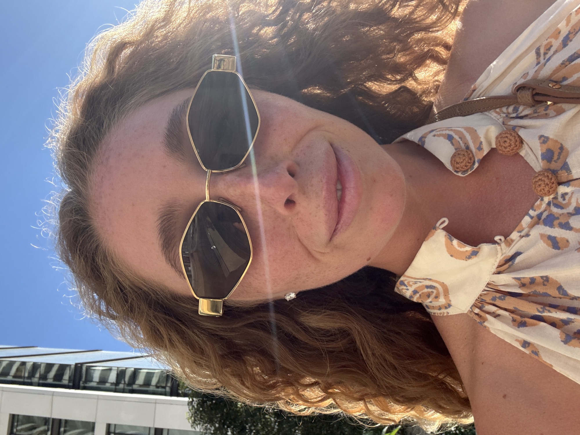A woman with curly auburn hair and freckles is pictured outdoors in Tel Aviv, Israel, in front of a modern building under a clear blue sky. She is wearing sunglasses with gold frames and a patterned halter top. Sunlight creates a lens flare across the image.