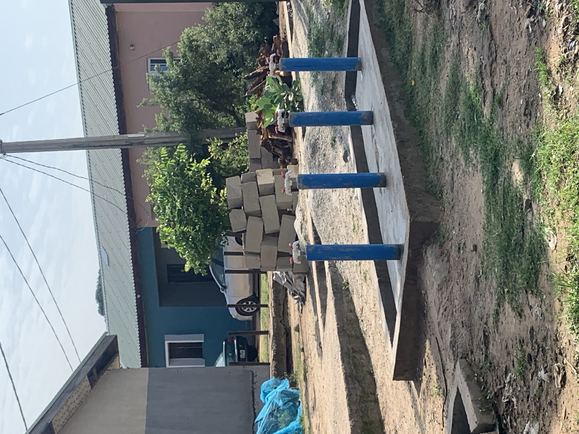 The image shows an outdoor scene with several buildings and a water standpipe. In the foreground, a concrete platform holds four blue water pipes with white and red spigots. The pipes are arranged in a row. Behind the pipes, there is a pile of concrete blocks and some debris. To the left, a blue tarp covers some items. In the middle ground, two cars are parked near a teal-colored building. Further back, a pink building is visible. The environment appears to be a mix of construction and residential areas, with dirt and grass covering the ground. A wooden utility pole with overhead wires is also present. The sky is visible in the background, with some clouds.