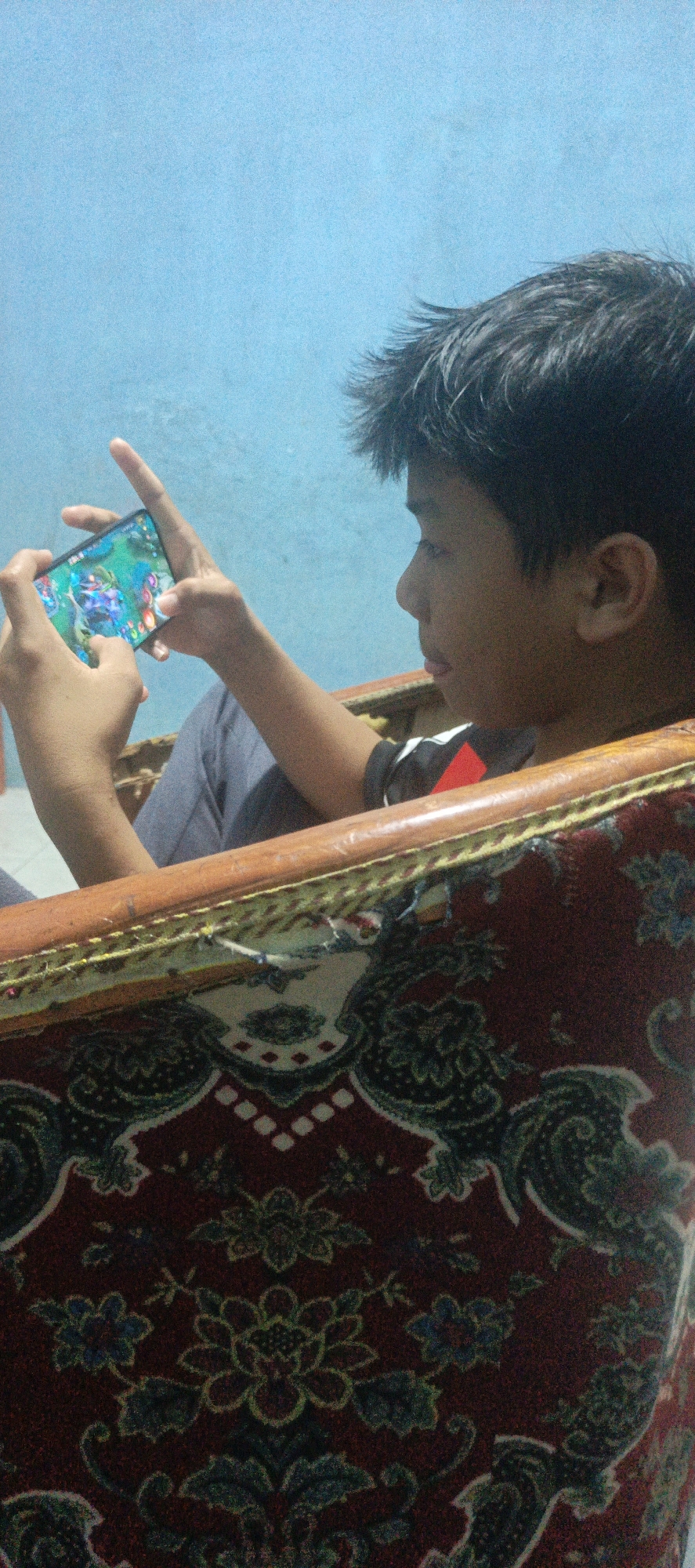 A young boy, appearing to be of Indonesian descent, is sitting in a chair and playing a video game on his smartphone. He is holding the phone horizontally with both hands, and his gaze is directed at the screen. He is wearing a dark t-shirt with red and white accents. The boy is seated in a chair that has a red, patterned fabric upholstery. The background consists of a light blue wall. The image is taken from a side profile, focusing on the boy and the immediate surroundings of the chair. The lighting appears to be from an overhead source. The image does not provide enough information to determine the specific location within the City of Pematang Siantar, Indonesia.