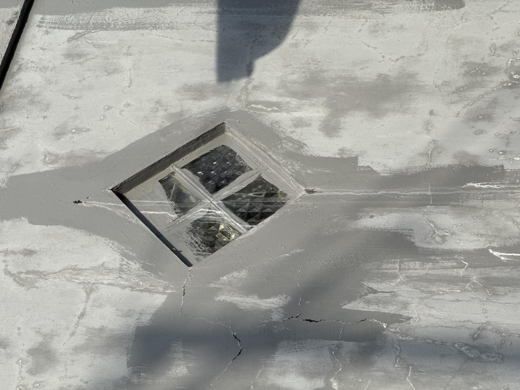 A diamond-shaped window with textured glass panes is set into a rough, gray stucco wall. The wall exhibits signs of age and weathering, with visible cracks, water stains, and patches of lighter and darker gray paint, suggesting a recent repair or application of a sealant. The window is framed by a slightly raised border, also finished in gray. Sunlight casts a shadow of an unseen object onto the right side of the wall. The scene appears to be an exterior wall of a building, likely residential or commercial, in Sukawati, Indonesia. There are no people or active events depicted in the image.