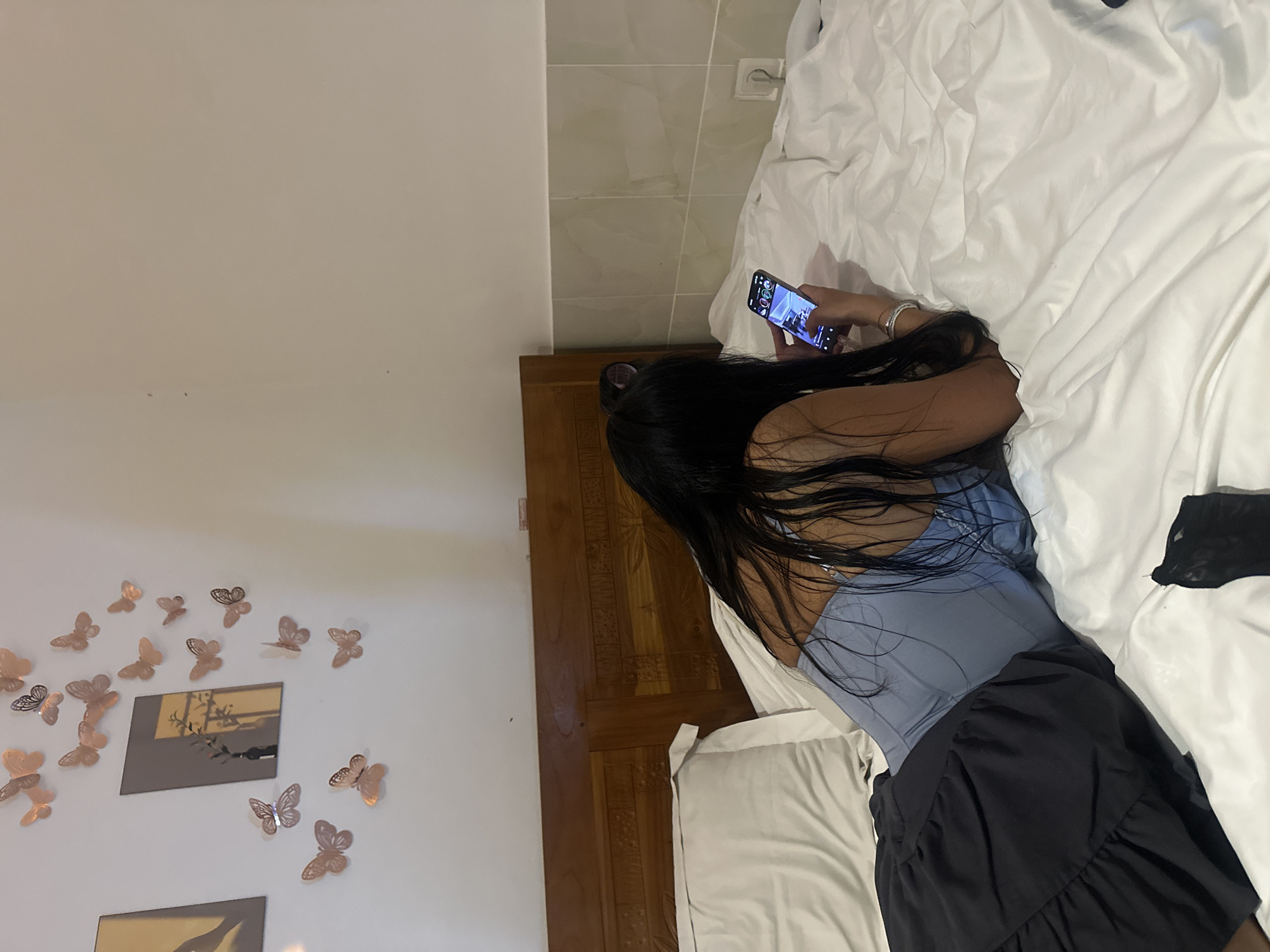 An indoor shot shows a person lying face down on a bed, absorbed in their smartphone. The image is oriented with the person's head towards the bottom right and their feet towards the top left when viewed upright.
The subject has long, dark hair and appears to be wearing a light blue, possibly corset-style, top and a dark-colored skirt with ruffles. Their right arm is bent, holding a smartphone with a brightly lit screen. The phone screen displays what looks like a social media or video application, showing circular profile icons, numbers like "43" and "4," and some indistinct text and interface elements. A play button icon is also visible.
The bed features white sheets and pillows. A dark piece of fabric, possibly another article of clothing, lies crumpled on the bed near the person's lower body. The bed frame, visible on the left side of the person, is made of dark wood and features intricate carved details.
The wall behind the bed (to the right of the person's head) is tiled with light-colored, possibly grey or beige, rectangular tiles. A white electrical outlet is visible on this wall. The wall on the far left (behind the person's back) is a plain, light color and is adorned with numerous metallic, butterfly-shaped wall decorations. Below these, two square or rectangular framed items, possibly mirrors or pictures with abstract designs, are also visible.
The scene depicts a moment of leisure or relaxation, likely in a bedroom or hotel room. Given the context of North Kuta, Indonesia, the wooden carvings on the bed frame might reflect local craftsmanship. The time of day appears to be evening or night, suggested by the reliance on the phone's bright screen for illumination.