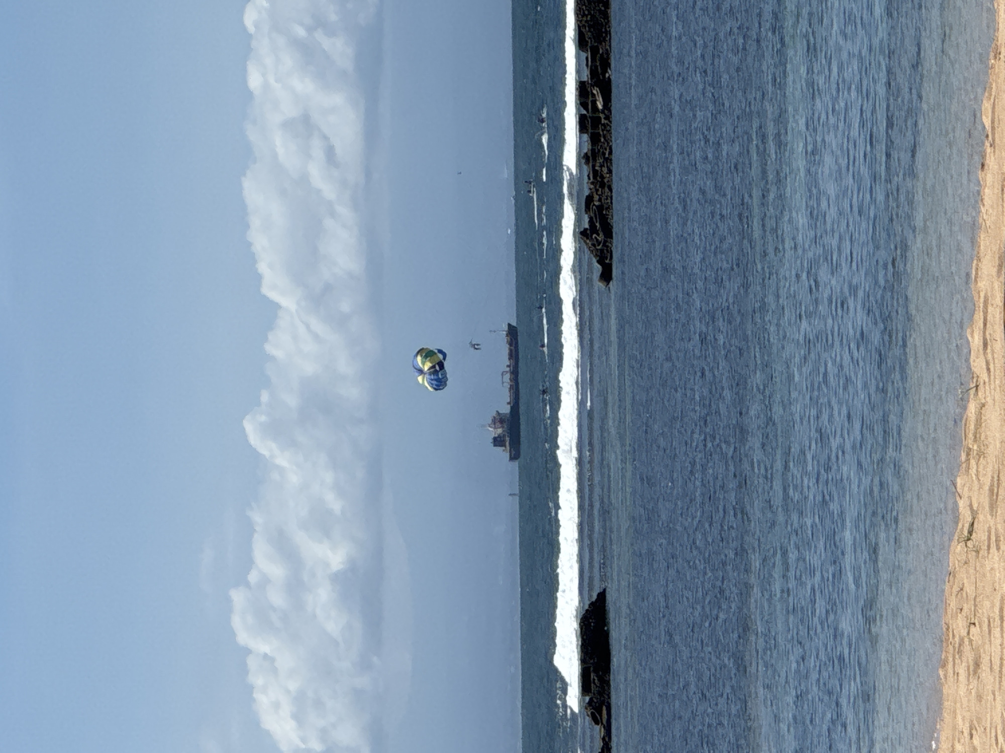 The image shows a beach scene in Kuta Selatan, Indonesia. On the right side of the frame is a sandy beach, and next to it is the ocean. Waves can be seen breaking along the shoreline. In the distance, a cargo ship is visible on the horizon. A parasailer is in the air, being pulled by a boat. The sky is light blue with large, fluffy white clouds on the left. A breakwater or pier, built from large rocks, is partially visible between the ocean and the beach.