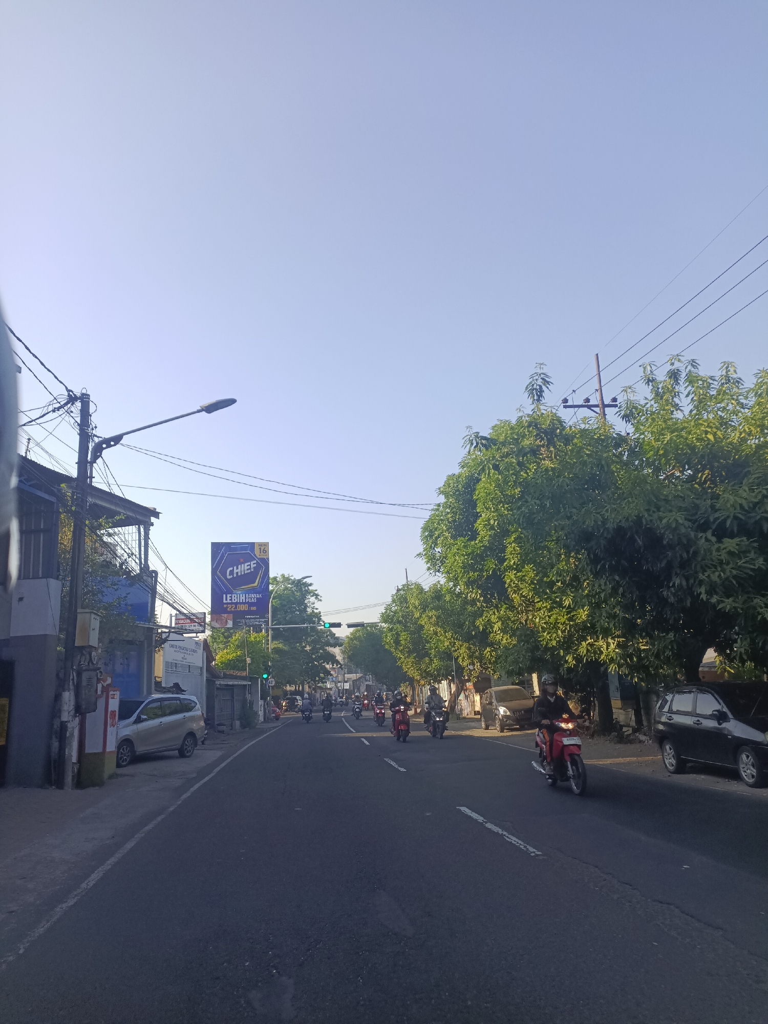 The image depicts a street scene in the city of Unknown, Unknown, likely during the daytime with clear skies. A paved road with a white dividing line runs through the center of the frame. Several motorcycles are in motion on the road, with riders visible on many of them. Some cars are parked along the side of the road, and others are also driving.
On the left side of the road, there is a building with a blue facade and a balcony. Utility poles with numerous wires are present throughout the scene, extending across the road and near the buildings. A large billboard advertising "CHIEF" with Indonesian text is prominently displayed on the left side of the road. Trees with dense green foliage line the right side of the street, creating a canopy effect in some areas. Traffic lights are visible further down the road. The overall environment suggests a bustling urban or suburban setting with active transportation.