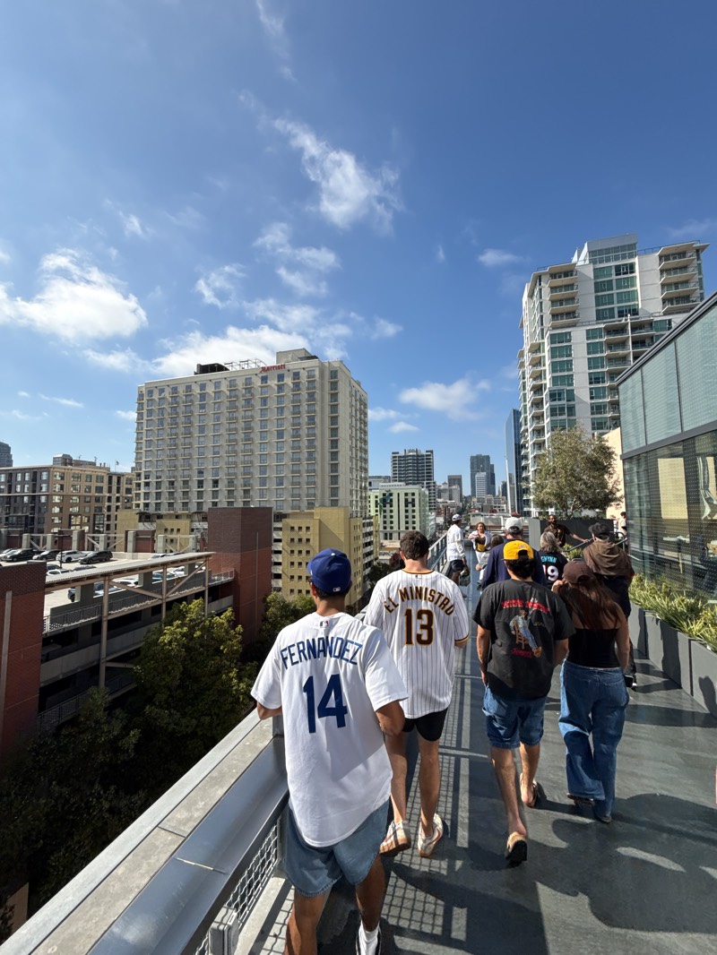 A full-shot photograph shows people walking on a pathway in downtown San Diego, California. The figures are walking away from the viewer, and consist of a group of adults and children dressed in casual clothing. The man closest to the camera is wearing a white shirt with a blue number 14 on the back. Beside him, a person is wearing a San Diego Padres baseball jersey with the number 13 on the back. Behind them are more people walking, with two in focus. One person is wearing a black t-shirt and blue shorts, while another is wearing a black tank top and blue jeans. Tall buildings are in the background and show an urban city. The sky is blue with white clouds. A tall parking structure is visible in the background to the left.