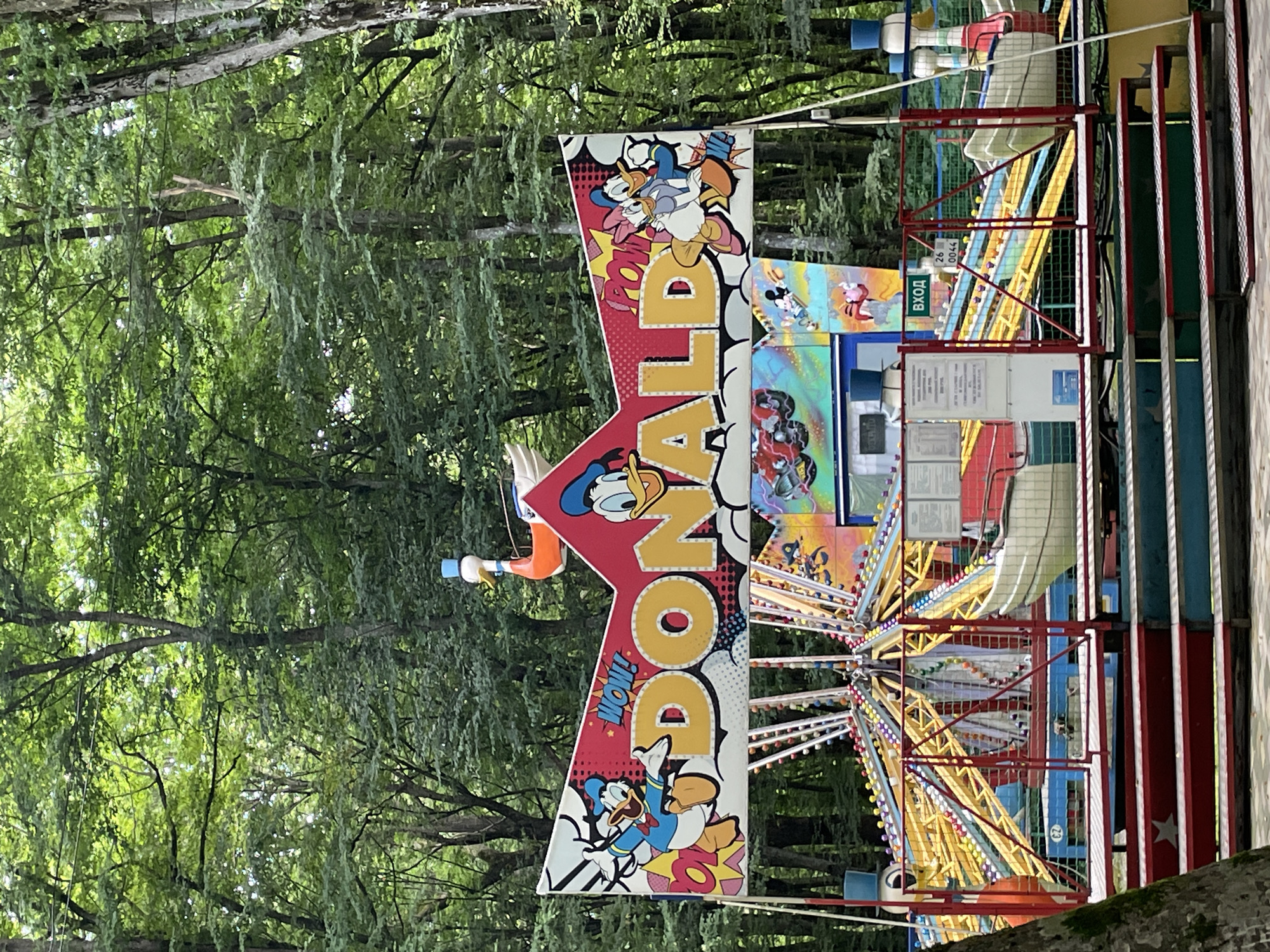 The image shows an amusement park ride with a sign that reads "DONALD" in large yellow letters, surrounded by cartoon characters from the Donald Duck franchise. The ride features colorful metal structures and seating areas. In the background, there are dense green trees, suggesting an outdoor, possibly wooded, location. A sign with Russian text, "ВХОД" (entrance), is visible. The overall scene depicts a children's attraction within an amusement park. The location of this specific amusement park ride within Stavropol, Russia, is not discernible from the provided image.