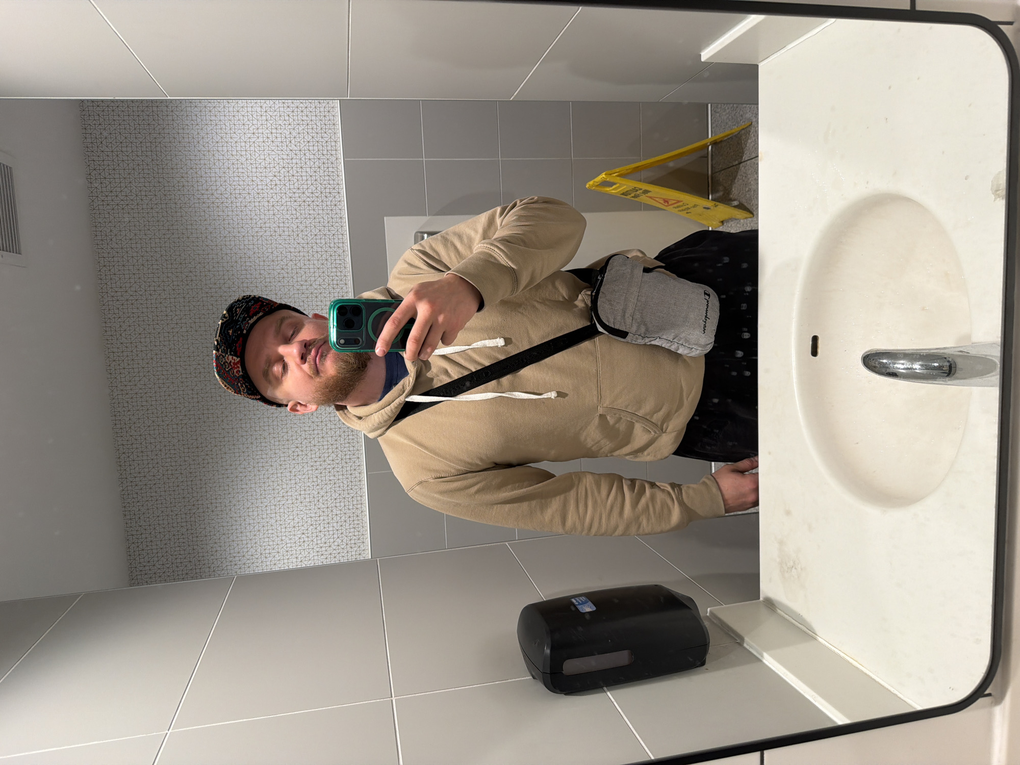 A mirror selfie in a public restroom. The person taking the photo is wearing a patterned cap, a tan hoodie, and a gray messenger bag with a black strap. They are holding a green smartphone. The restroom has gray and white tiled walls. A white sink with a chrome faucet is visible in the foreground. A black soap dispenser is on the floor. A yellow wet floor sign is in the background. It is not possible to determine the city of Chicago, Illinois as the exact location.