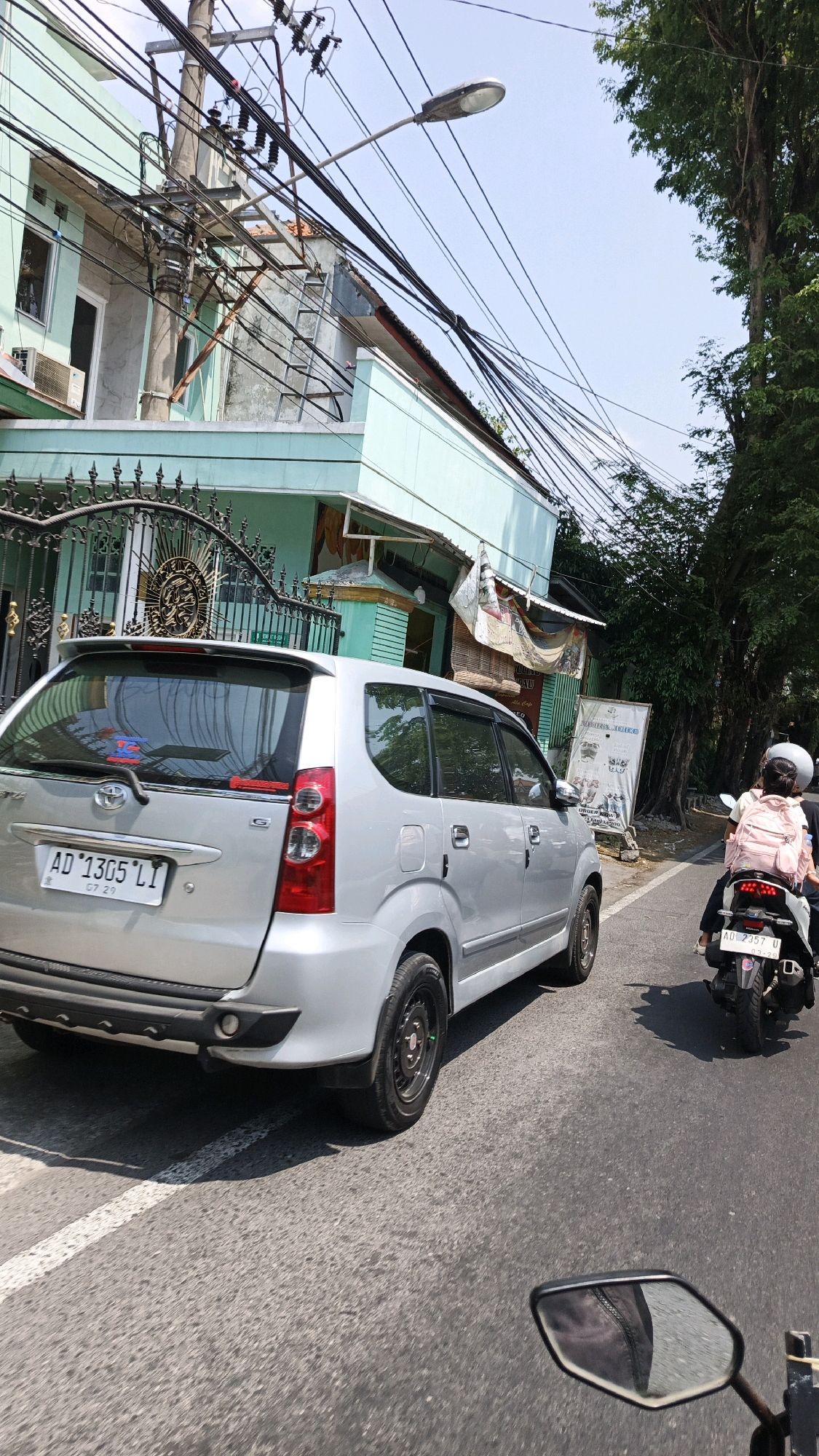 The image shows a street scene in Surakarta, Indonesia. In the foreground, a silver minivan with license plate "AD 1305 LI" is parked on the right side of the road. A motorcycle with a rider wearing a white helmet and a pink backpack is passing the minivan on the right. The motorcycle has license plate "AD 2357 U". The street is paved and appears to be in a residential area, with houses and trees lining the sides. A utility pole with numerous electrical wires is visible in the background, along with a light fixture. The sky is clear and blue.