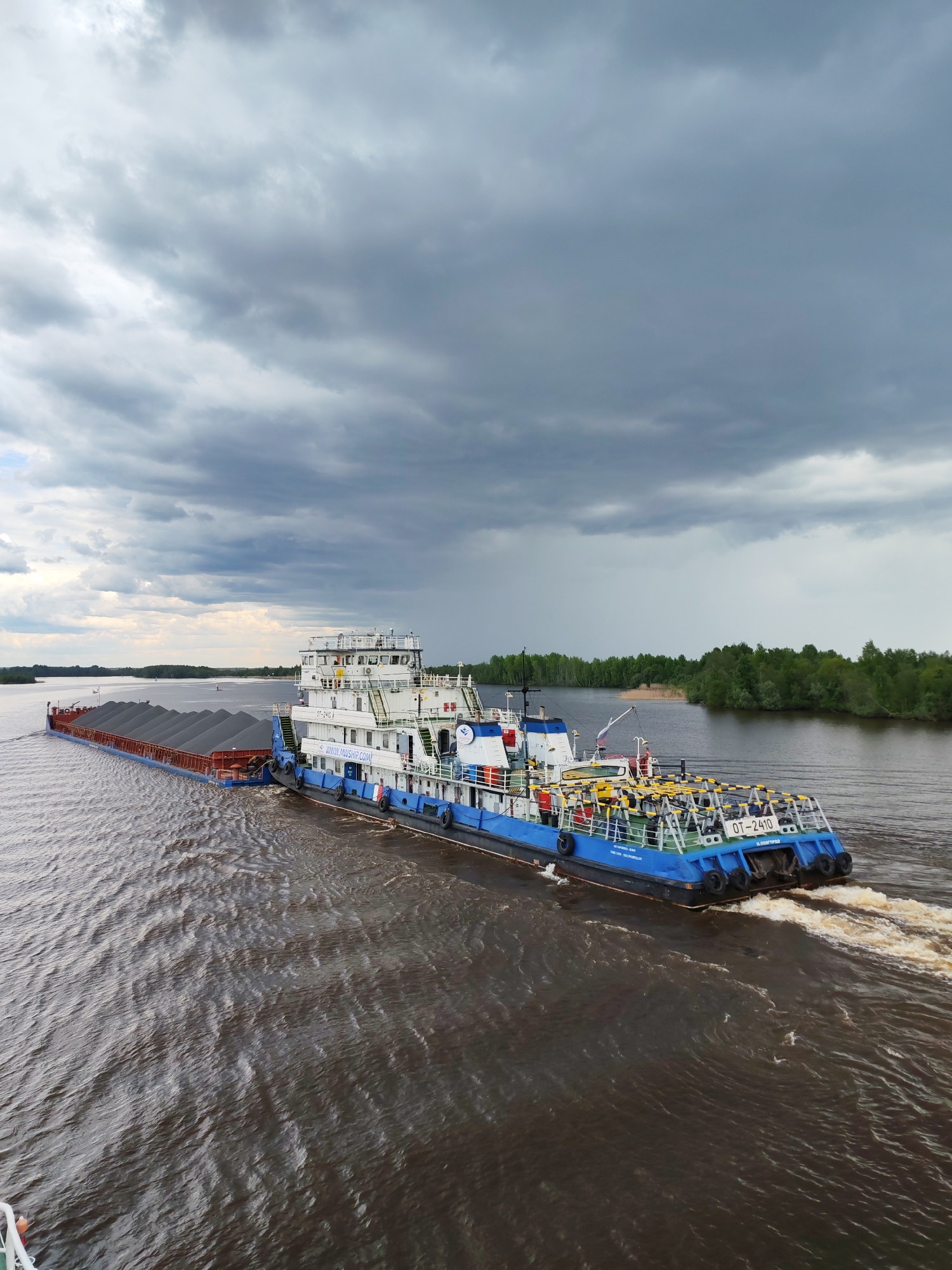 A river scene in what appears to be a cloudy environment. A blue and white tugboat, "OT-2410," is pushing a barge laden with dark material, possibly coal or gravel. The scene is likely near a riverbank, as indicated by the tree line visible on the right side of the frame. No people are clearly visible. Given the location provided, this is presumably happening in or near Kargulino, Russia. The water is dark and disturbed by the movement of the vessels.