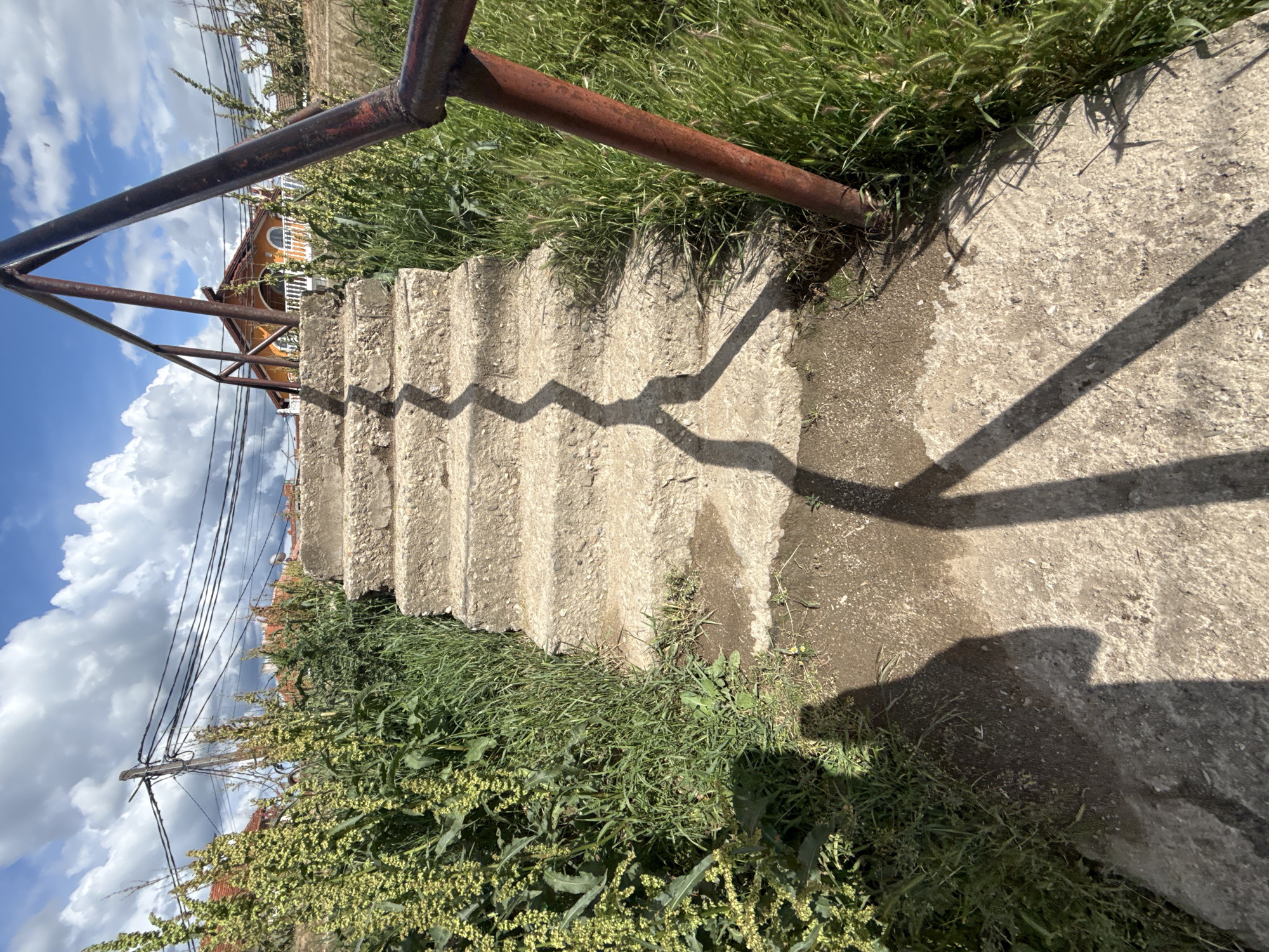 The image displays an outdoor scene featuring a set of concrete stairs leading upwards. A dark brown, somewhat rusty metal railing runs alongside the left side of the stairs, extending into the foreground. Lush green grass and weeds grow abundantly on both sides of the staircase and railing.
Strong, bright sunlight illuminates the scene, casting distinct, jagged shadows of the railing across the concrete steps and the ground to the right. This suggests it is midday or early afternoon. The ground next to the stairs on the right appears to be a mix of dirt and concrete, with some damp or darker patches.
In the background, visible through and above the railing, is part of a house or building. It has an orange or ochre-colored facade, with a white railing on a balcony and an arched window. Above the house, several power lines or utility wires stretch across the sky. The sky itself is bright blue with scattered, fluffy white cumulus clouds, indicating fair weather. There is no discernible human activity or visible text in the image. The setting appears to be a residential or semi-rural outdoor area.