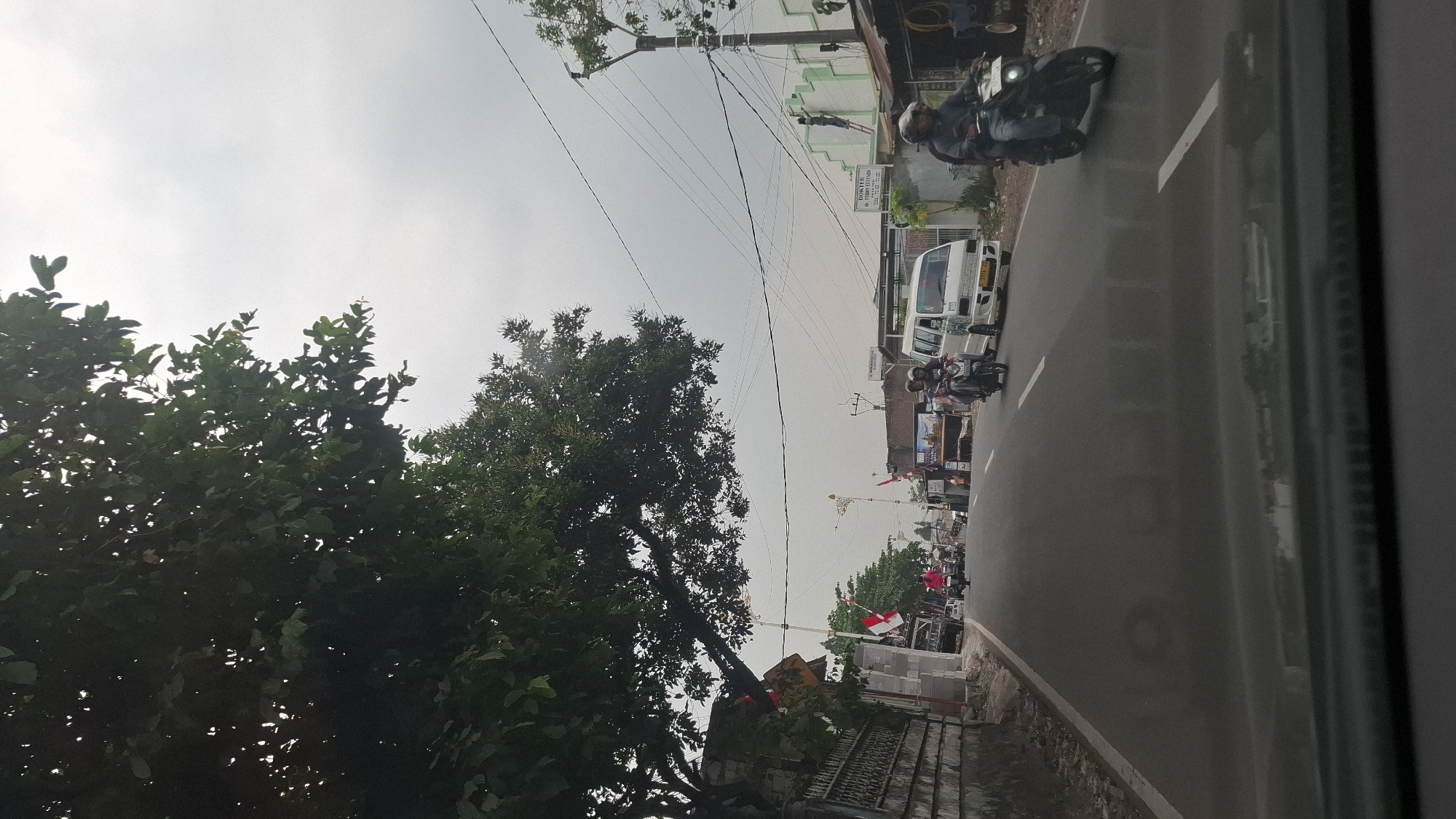 The image shows a street scene in Cilaku, Indonesia, with vehicles and pedestrians on the road. The sky is overcast. On the left side of the road, there is a stone wall with a large tree providing shade. Several Indonesian flags are visible along the street. A white van and two motorcycles are driving down the road. On the right side of the road, there is a building with a sign that reads "DOKTER DR. FERRY ELINDO". A person is on a ladder on the side of this building. Another motorcycle is passing by on the right. The scene appears to be a typical street in a semi-urban area.