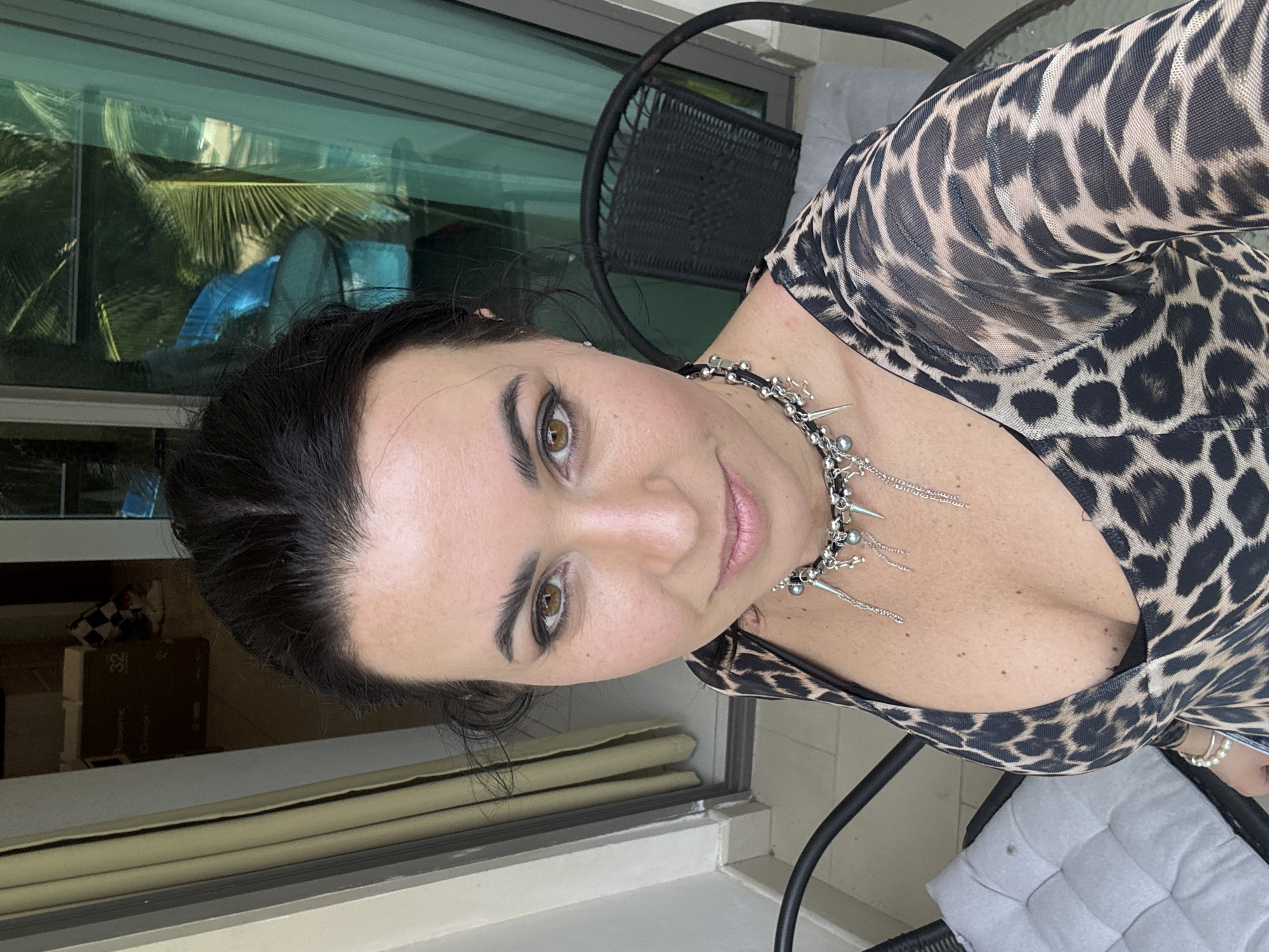 A woman poses for a selfie on a balcony. The woman has dark hair, fair skin, and brown eyes. She is wearing a black and tan leopard-print top and a silver necklace with dangling charms. Behind her is a sliding glass door that leads to an outdoor space. Through the glass, greenery and a blue object are visible. A black metal chair is also present in the scene. Some nondescript boxes are in the background, on the left. The location is likely Pattaya City, Thailand, based on the possible presence of tropical foliage.
