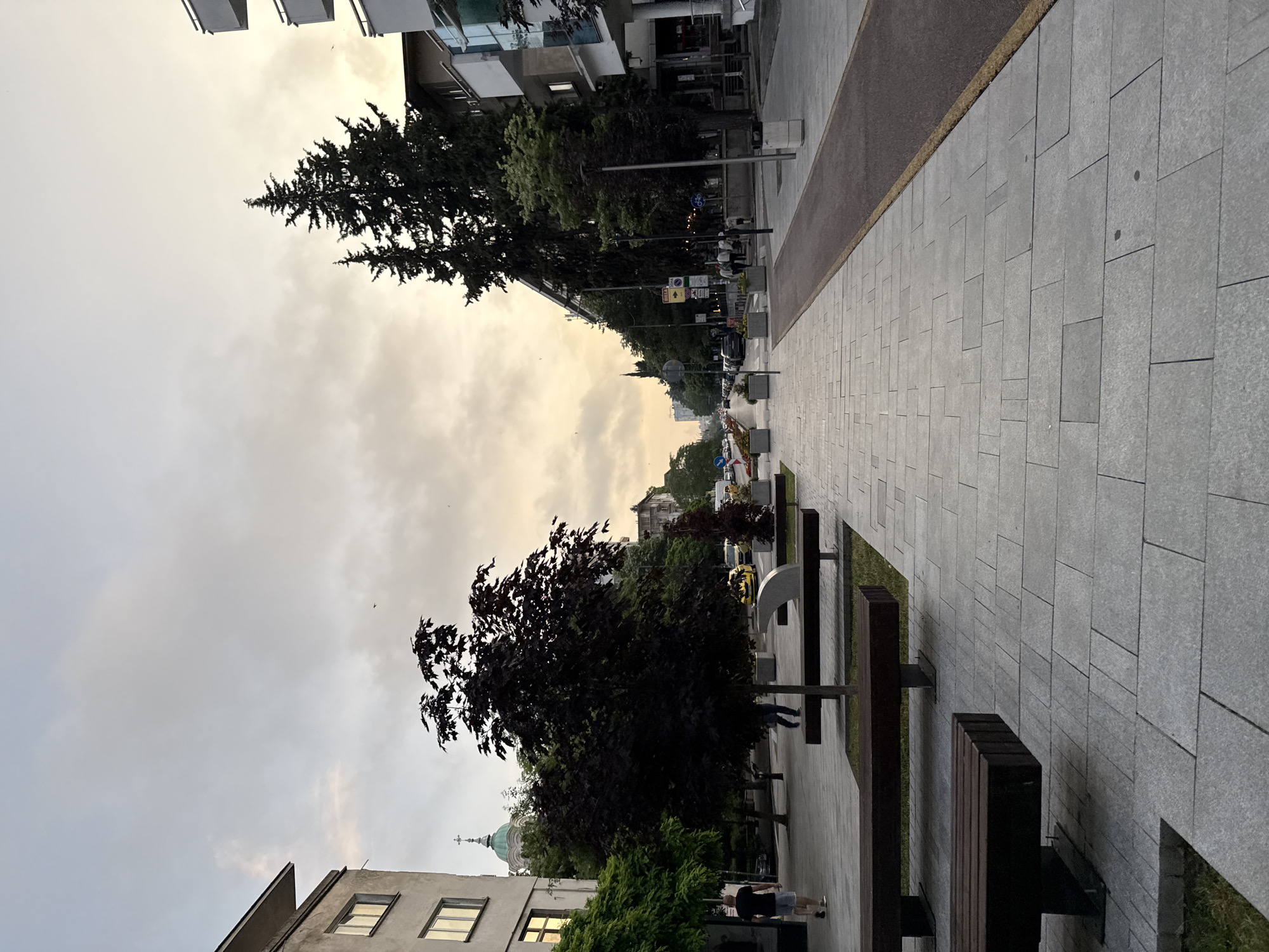 This media file captures an urban street scene in Stara Zagora, Bulgaria, during what appears to be late afternoon or early evening. The sky is overcast but shows patches of warm, golden light, suggesting the approach of dusk or sunset.
The scene features a wide, modern pedestrian walkway paved with grey rectangular and square tiles, stretching from the foreground into the distance on the right. Parallel to this path is a darker asphalt road. Several dark, modern wooden benches are arranged along the pedestrian area, with one person walking near a bench in the midground.
To the left of the walkway, the street is lined with mature trees, some with dark, dense foliage, and further down, a tall, conical evergreen tree stands out. Behind the trees, various multi-story buildings with balconies and windows are visible, indicating a residential or commercial area. A notable landmark in the mid-left is the distinctive green dome topped with a white cross, belonging to a church, rising above the surrounding buildings.
Further down the street, more parked cars and a few distant pedestrians can be seen. Several street signs are visible, including a blue circular sign with a white bicycle symbol, indicating a bicycle lane or route. However, specific text on these signs is not clearly legible due to distance. The overall atmosphere is tranquil, portraying everyday life in a European city.