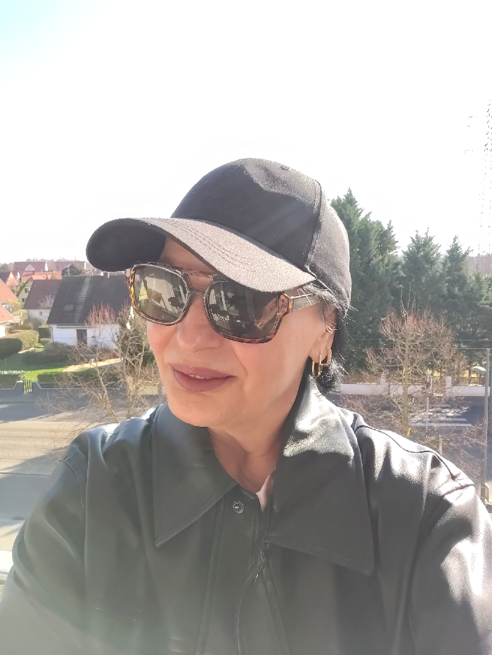 A woman is depicted from the chest up, positioned outdoors on a clear, sunny day in Ostwald, France, possibly on a balcony or elevated platform overlooking a residential area. She is wearing a black baseball cap, tortoiseshell-patterned oversized sunglasses, and a black leather-look jacket with a prominent collar and visible snap fasteners. Her lips are made up with a subtle reddish-brown lipstick, and she has dark hair visible beneath the cap and is wearing small gold hoop earrings on her visible right ear. The woman is looking slightly to her left, with a neutral facial expression. The background features a suburban landscape with several light-colored residential houses topped with red-tiled roofs, a paved street, and various trees, some bare-branched, and a cluster of taller evergreen trees further in the distance under a bright, largely cloudless sky.