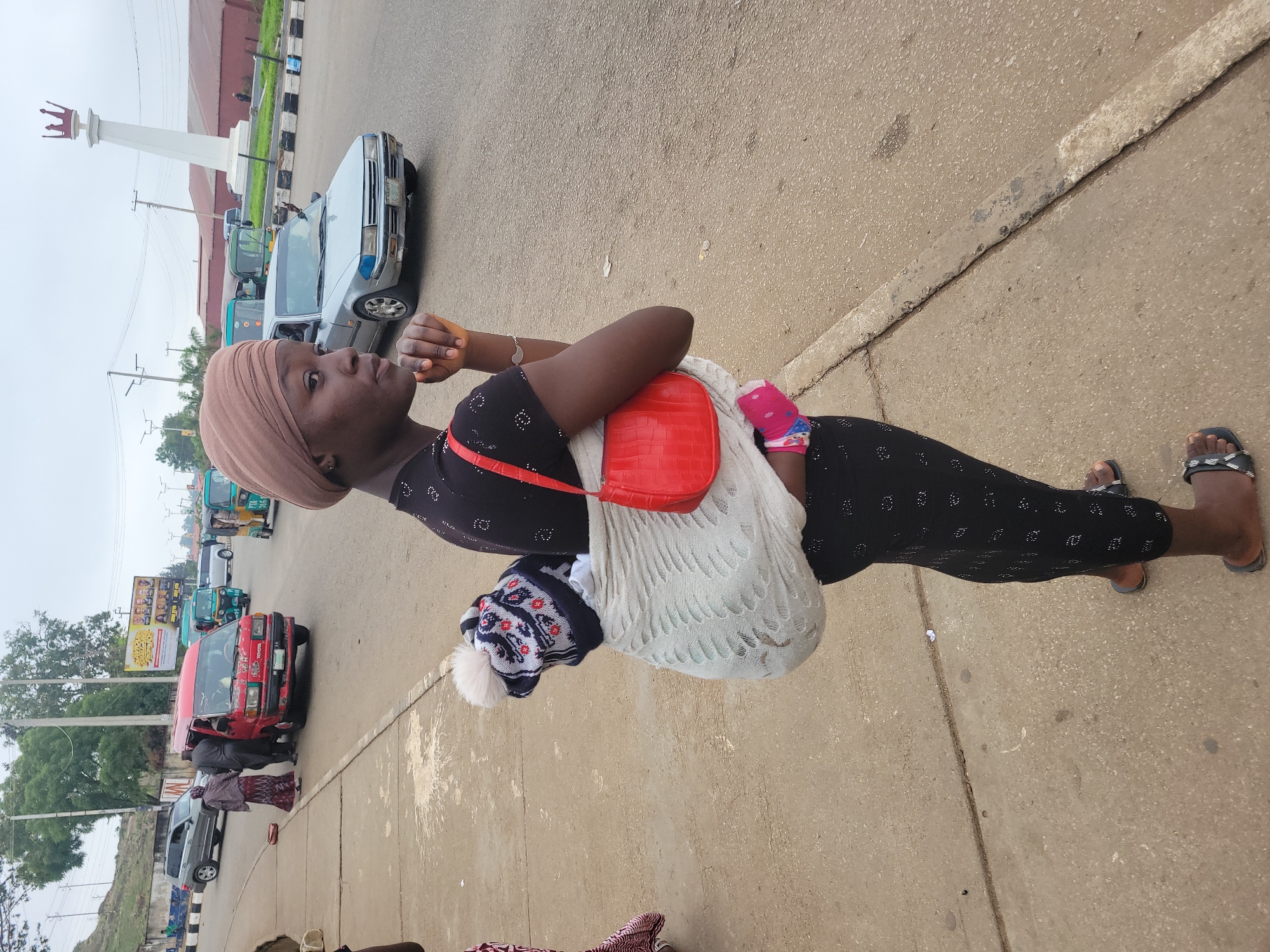 A woman stands on a sidewalk in Jos, Nigeria, facing away from the camera. She is carrying a baby wrapped in a white cloth on her back. The baby is wearing a patterned hat. The woman has a red purse slung over her shoulder. She is wearing a black patterned dress and black sandals. In the background, there are cars and other people on the street. There is also a tall white monument with a crown on top. The sky is overcast.