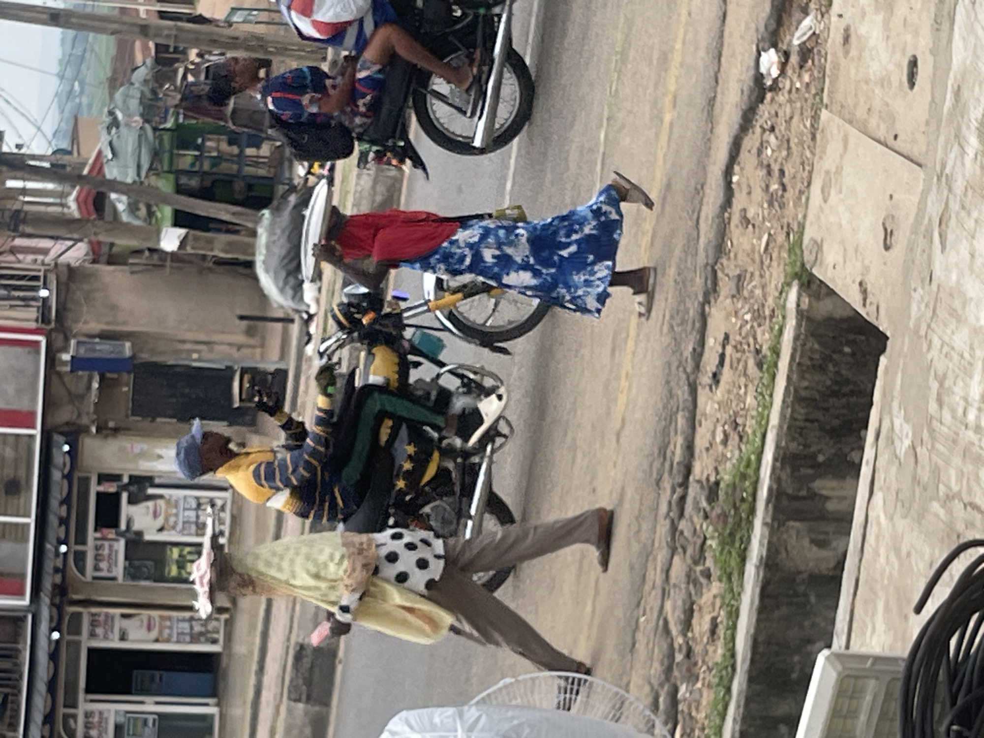 In this street scene, three individuals are captured in motion. A person wearing a yellow and blue striped shirt, dark pants, and a blue cap is riding a motorcycle. Beside them, a person in a light yellow head covering and a black and white polka dot top walks, carrying an object on their head and a pink item in their hand. To the right, a person in a blue and white tie-dye dress and a red head covering walks and carries a white tray on their head. Behind them, another motorcycle is visible with a person in a blue and patterned dress riding on the back. The environment appears to be a street with buildings and a paved road, with a concrete drainage channel in the foreground.