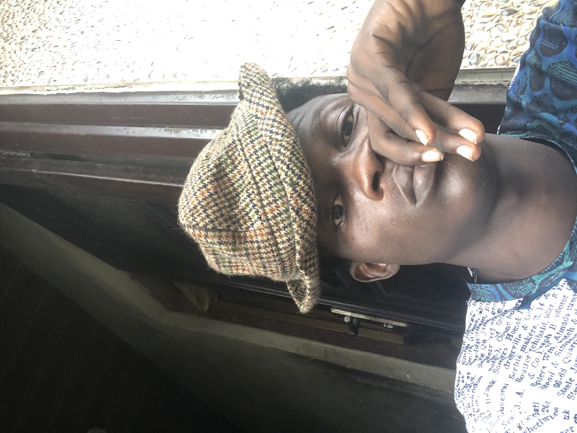 A close-up, head-and-shoulders selfie of a young Black male wearing a tweed flat cap and a patterned shirt is captured in a dimly lit, outdoor setting in Ajah, Nigeria. The subject's face is partially obscured by his hand, which is resting on his mouth, with his fingers extended. He is looking directly at the camera with a neutral expression. The background consists of a wooden door frame and a speckled wall. The lighting is dim, casting shadows across the subject's face and the environment. The shirt has a white base with dark blue and green lettering and patterns.