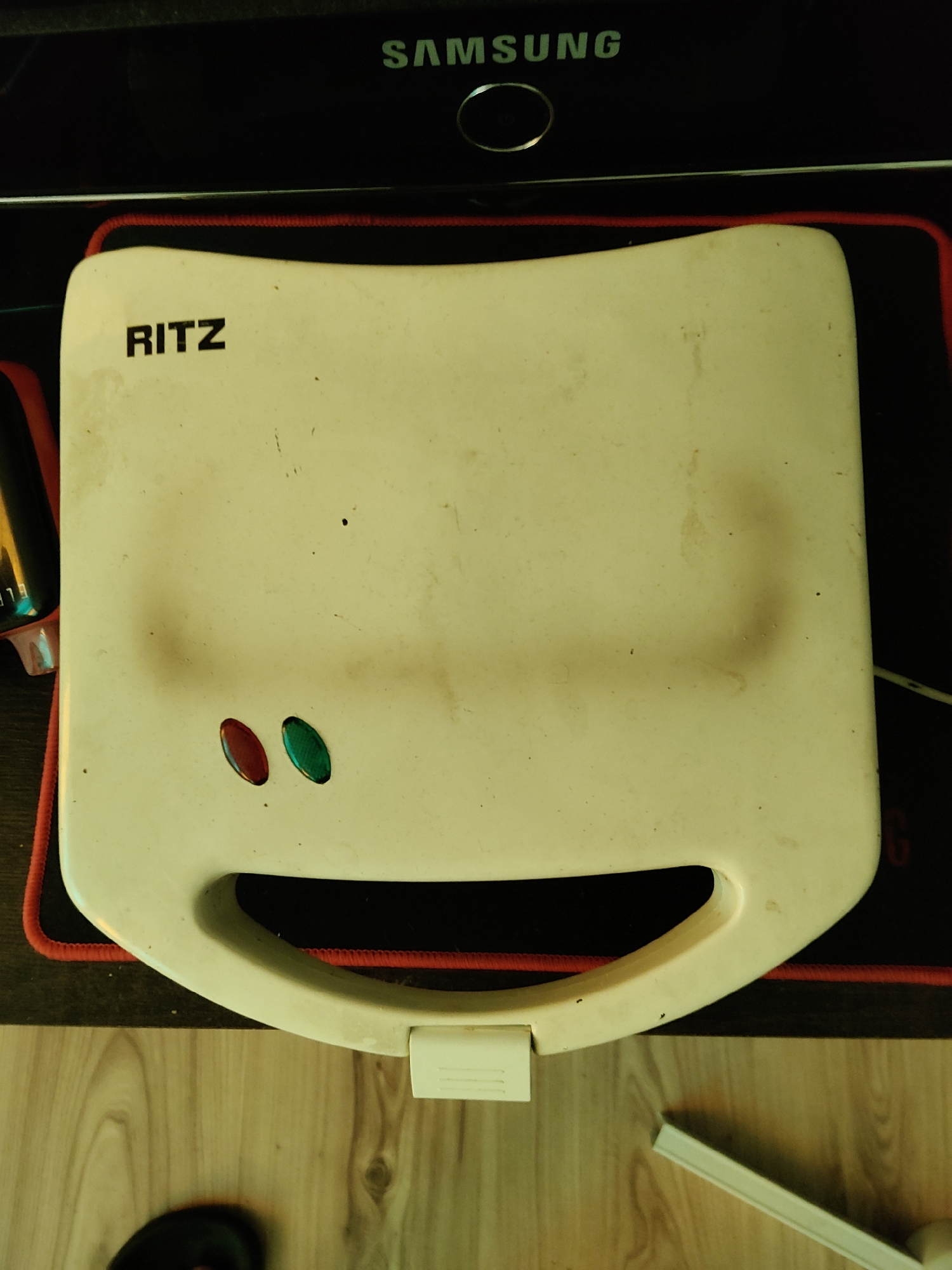 The image shows a close-up, top-down view of a white Ritz sandwich maker on a black mousepad with red trim. The sandwich maker is old and shows signs of use, with a few dark specks and a faint stain on its surface. To the left of the sandwich maker, the corner of a black object with a yellow reflective surface is visible. Above the sandwich maker, the word "SAMSUNG" is visible in green lettering on a black background. The background also shows a portion of a wooden surface. The scene does not provide any information about the city of Płock, Poland.