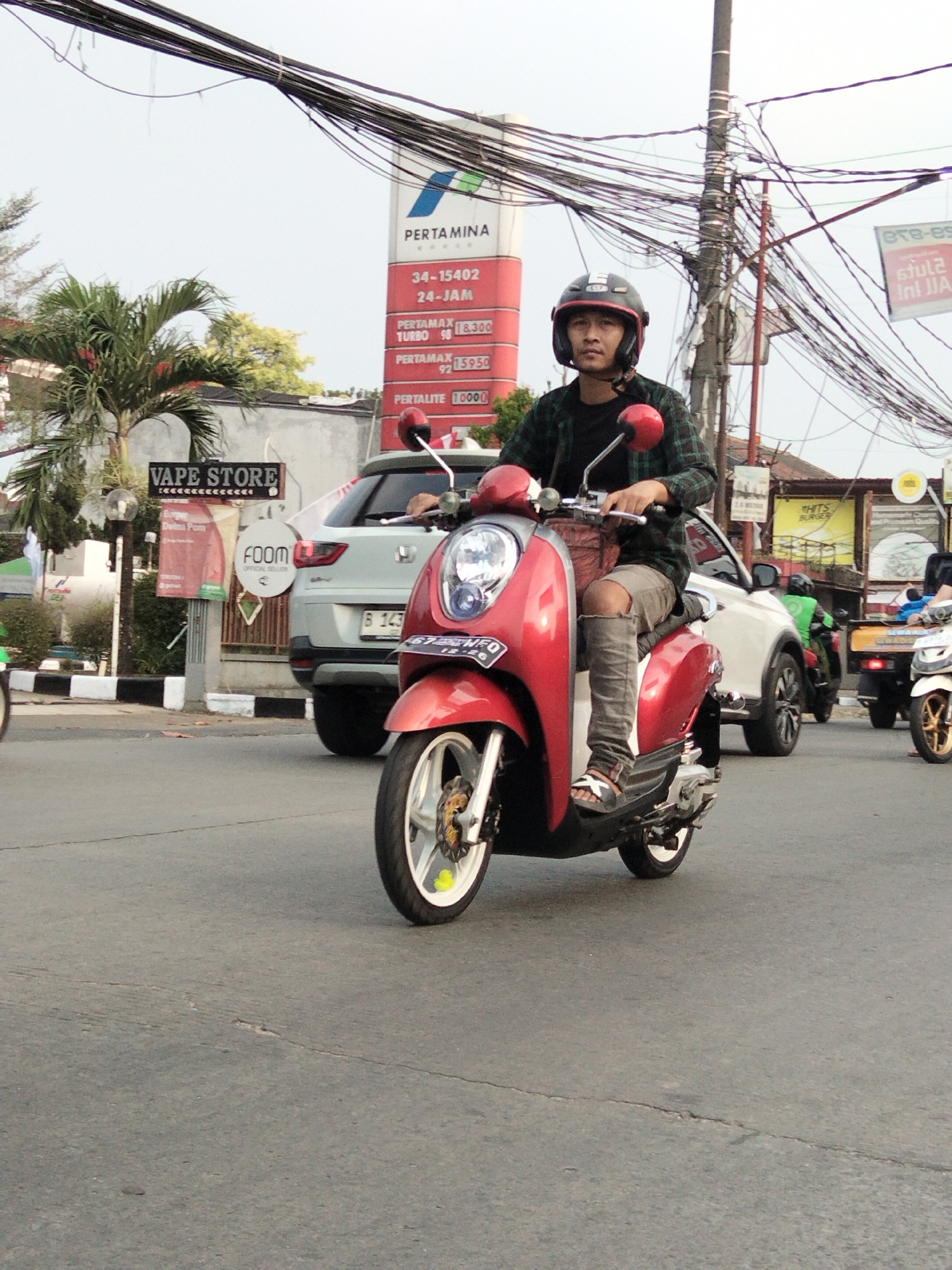 The image depicts a street scene in South Tangerang, Indonesia, on an overcast day. In the foreground, a man wearing a helmet and a plaid shirt is riding a red scooter. He is wearing ripped grey pants and sandals. Behind him, a white SUV is visible, and further back, other vehicles and buildings line the street. A prominent red and white Pertamina gas station sign is visible in the background, displaying fuel prices and operating hours. The street is paved, and there are numerous utility poles with tangled wires overhead. Signs for a "Vape Store" and "FOOM Official Seller" are also present, suggesting a commercial area. The overall atmosphere is that of a typical urban street with active traffic.