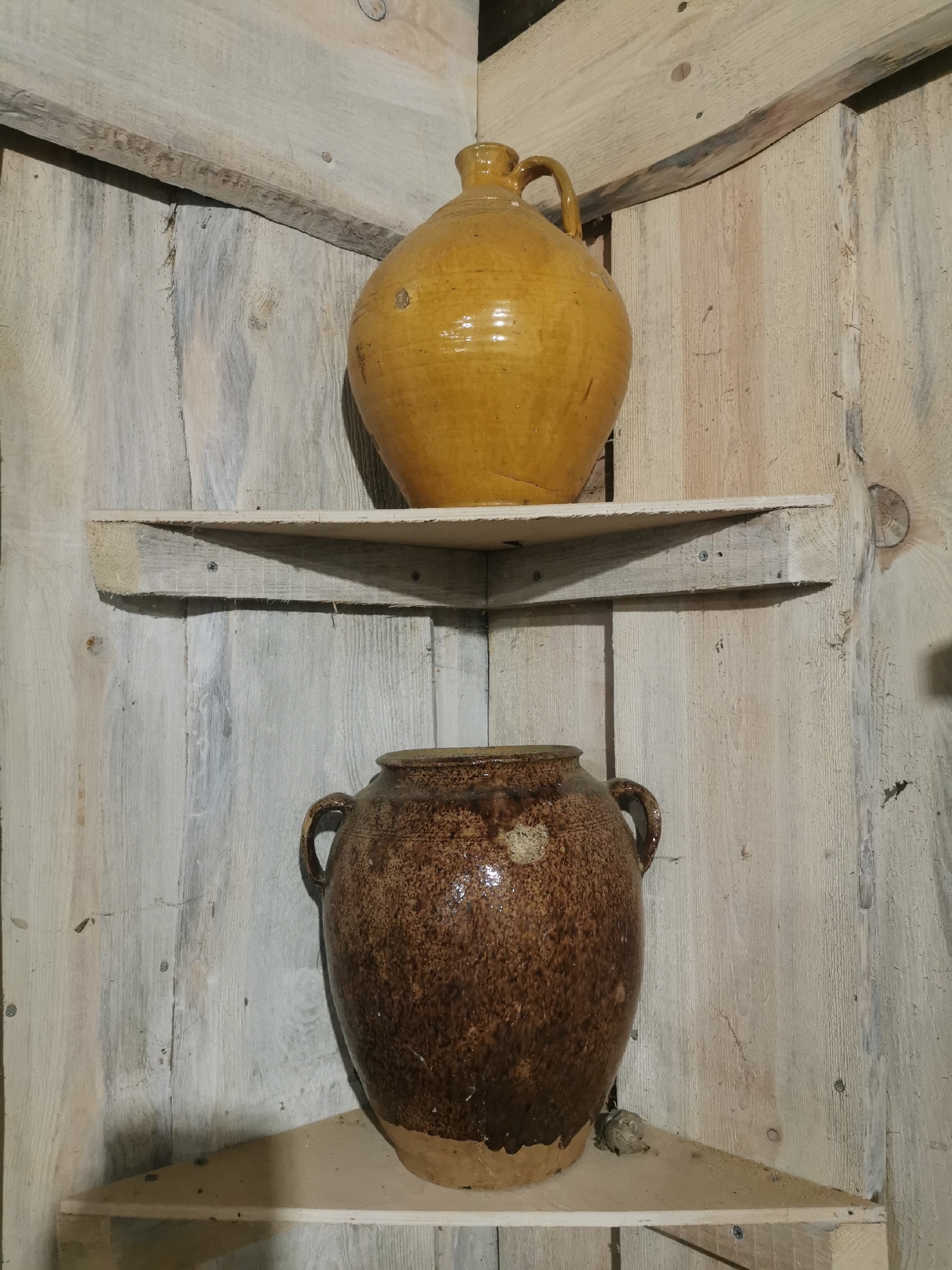Two earthenware jars are displayed on wooden shelves in a corner of a room. The upper shelf holds a large, round, yellow-glazed jug with a handle. The lower shelf holds a darker brown, speckled jar with two handles. The walls are made of weathered wooden planks.