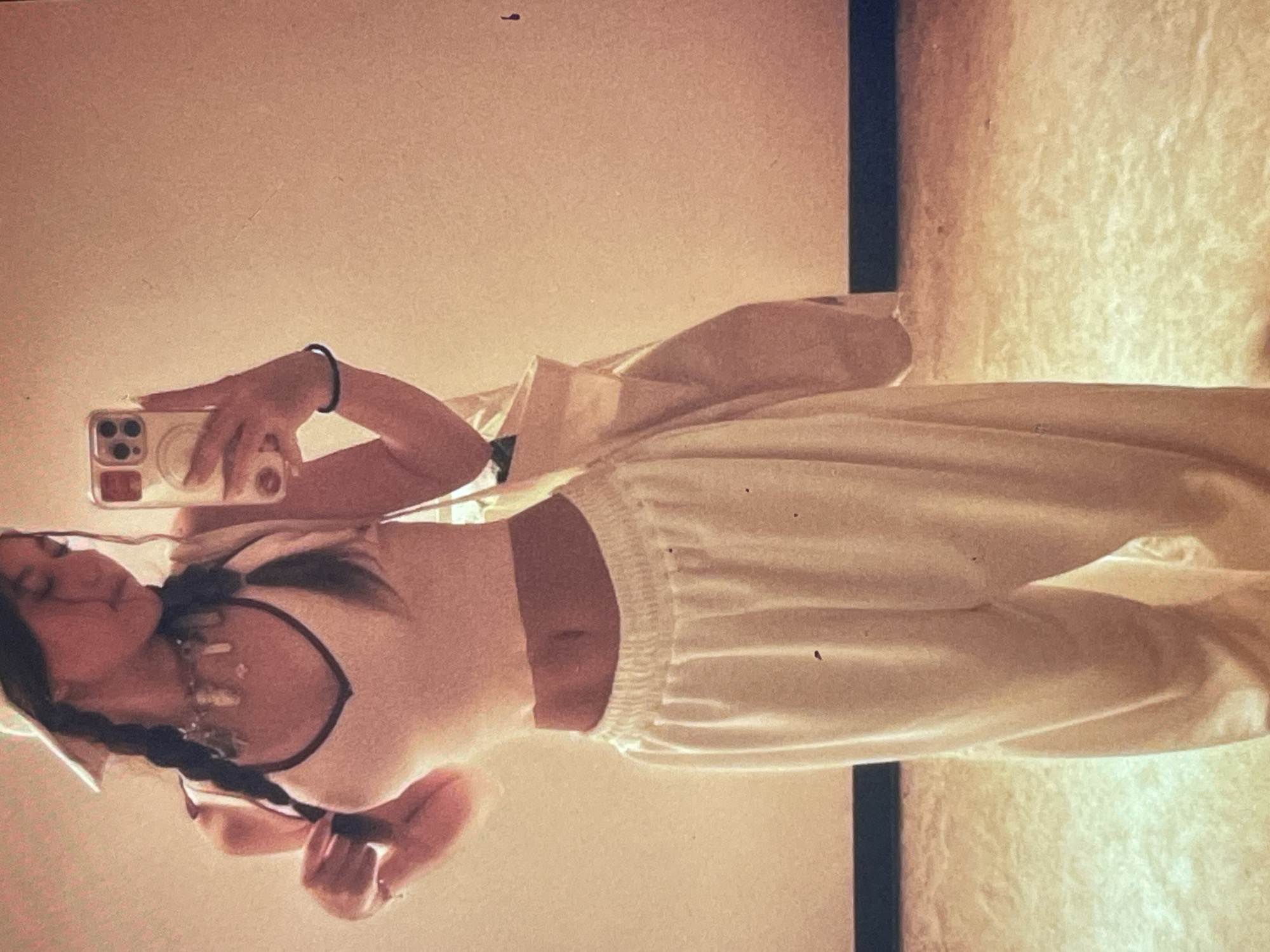 This media file captures a young woman taking a mirror selfie indoors in Poltava, Ukraine. The image is oriented vertically, depicting the woman from just above her head to her feet, reflected in a large mirror.
The main subject is a young woman with dark hair styled in two long braids. She is wearing a light-colored, possibly cream or off-white, sleeveless crop top with dark trim around the neckline, revealing her midriff. She pairs this with loose-fitting, light-colored baggy pants. On her right wrist, she wears a simple black bracelet or hair tie. Around her neck, multiple layered necklaces with what appear to be charms or small crystal pendants are visible.
The woman holds a light-colored smartphone, possibly an iPhone, in her right hand, positioned in front of her face as she captures her reflection. Her left hand is partially visible near her hip. A light-colored bag or pouch is draped over her left shoulder or arm.
The background shows a simple interior setting. The wall behind the woman is a solid, warm, light brown or peach color. To the right of the mirror, a darker, vertical element (possibly a doorframe or another piece of furniture) is visible. The floor area below the mirror appears to be a lighter, textured surface, possibly carpet or a rug. The lighting is soft and warm, suggesting an indoor setting with ambient light, though the exact time of day is not discernible. No visible text is present in the image.
