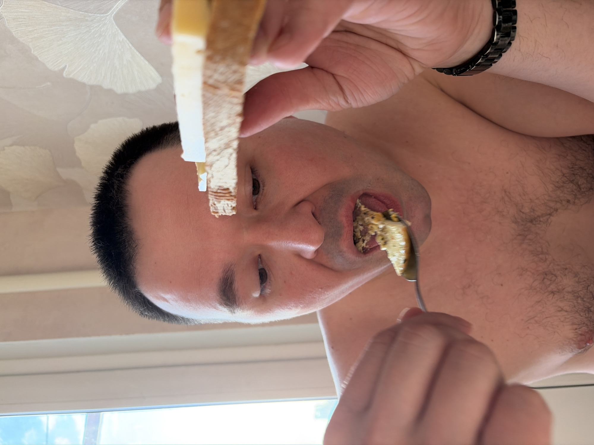 A medium shot depicts a fair-skinned, shirtless man with short, dark hair, eating with a spoon. He appears to be holding a piece of bread in his left hand. A black bracelet is visible on his left wrist. He is looking at the camera, and his mouth is open. The background includes a white ceiling decorated with a repeating ginkgo leaf pattern, and a window with a glimpse of a blue sky. The location is Izhevsk, Russia.