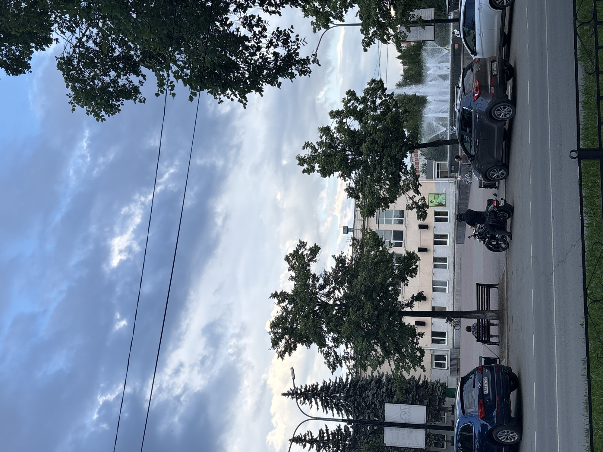 The image shows a street scene with a fountain in the background, suggesting a public park or square. The sky is overcast with dramatic clouds. On the right side of the frame, there is a road with parked cars and a motorcycle. A person is sitting on a bench under a tree, and another person is on the motorcycle. The building in the background appears to be a multi-story apartment complex. There is some visible text on signs, but it is difficult to read. The overall impression is a somewhat gloomy day in an urban or semi-urban setting. The presence of a fountain and trees indicates a public space. The time of day is unclear due to the overcast sky, but it appears to be daytime.
