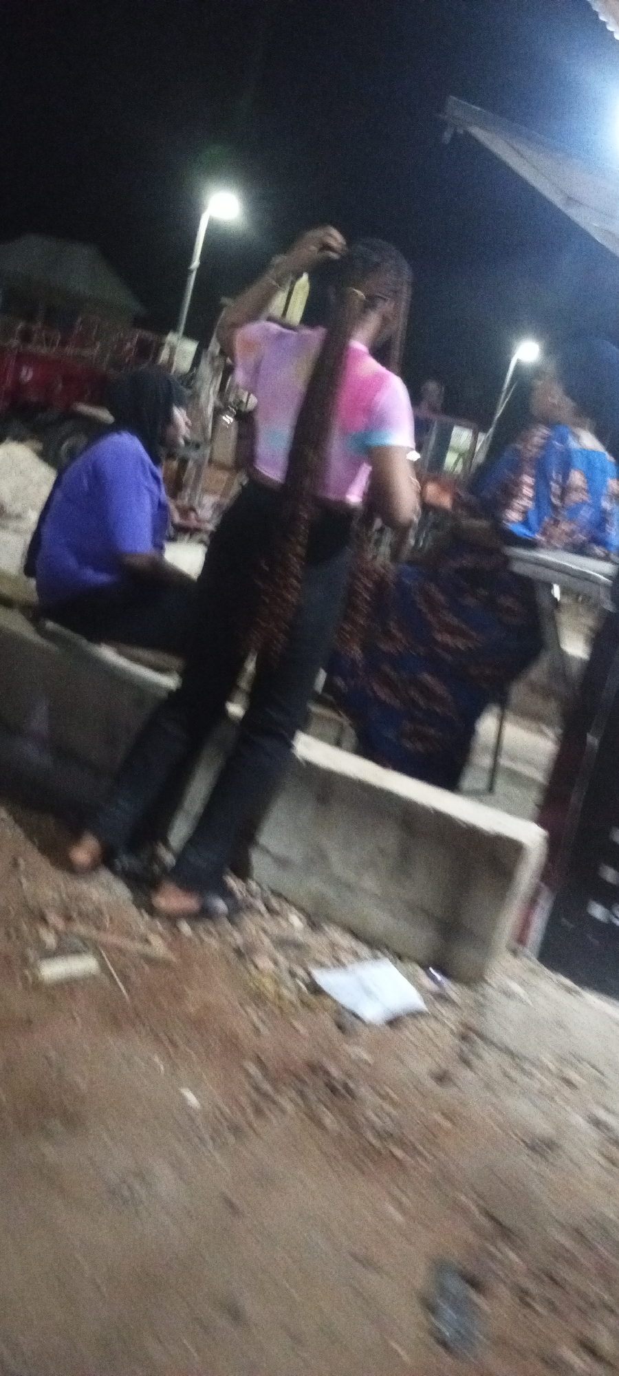 At night in Adankolo, Nigeria, a group of people are gathered outdoors. A person in a tie-dye t-shirt and black pants is having their hair braided. To the left, another person in a purple shirt and black pants sits on a concrete barrier. In the background, a person in a blue and patterned dress is visible. The scene appears to be in a dimly lit area with a concrete barrier and rough ground.