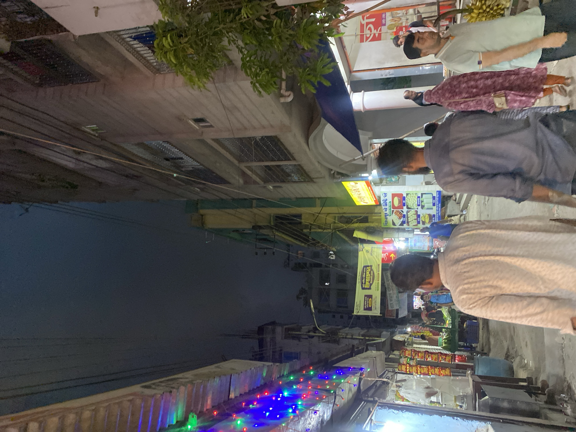 The image depicts an evening scene in Dhaka, Bangladesh. A street lined with buildings is visible, many displaying signs in Bengali. A canopy of colorful lights runs along a building's edge in the lower left. Several people are present, walking along the street. One individual is selling bananas from a cart. The overall atmosphere appears to be a typical evening street scene in a busy urban area.