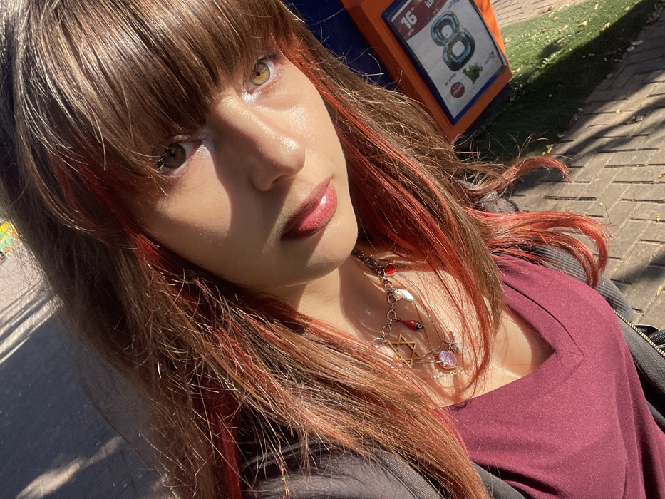 The image is a close-up, upward-angled selfie of a young female presenting individual outdoors in Raanana, Israel. The subject has medium-brown hair with prominent red streaks and bangs. Her eyes appear heterochromatic, with the left eye (viewer's left) being hazel/greenish-brown and the right eye (viewer's right) lighter, possibly amber, with a bright reflection. Her lips are glossy and full. She wears a deep maroon V-neck top and a dark jacket. A silver-colored chain necklace adorns her neck, featuring charms including a golden Star of David, small red teardrop shapes, a heart, a small evil eye bead, and another iridescent pendant. The lighting is strong, casting sunlight across her face and deep shadows on the ground. The background shows a paved sidewalk made of interlocking concrete pavers and a strip of green grass. To the right, a distinctive orange public utility or information box is visible, displaying white numerals "16" and "8", and Hebrew text "יוני" (June), confirming the Israeli context.