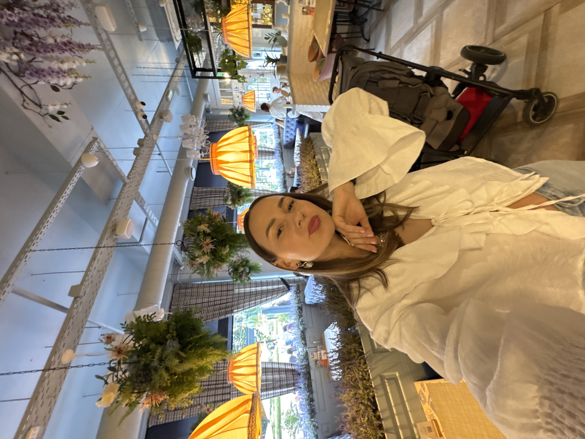 A medium-angle selfie depicts a woman indoors, apparently in a commercial space. She is positioned in the foreground, wearing a white, ruffled blouse, her hand near her face.
In the background, the space appears to be a restaurant or similar establishment. Visible are hanging floral decorations and pendant lamps, suggesting a carefully designed interior. Further into the space, tables and seating can be seen, with other patrons seated in the distance. A stroller is also nearby.
The location might be in Sochi, Russia, as the environment resembles locations of that area. The orientation of the image is at an angle, and the details are presented without any added subjective interpretation.